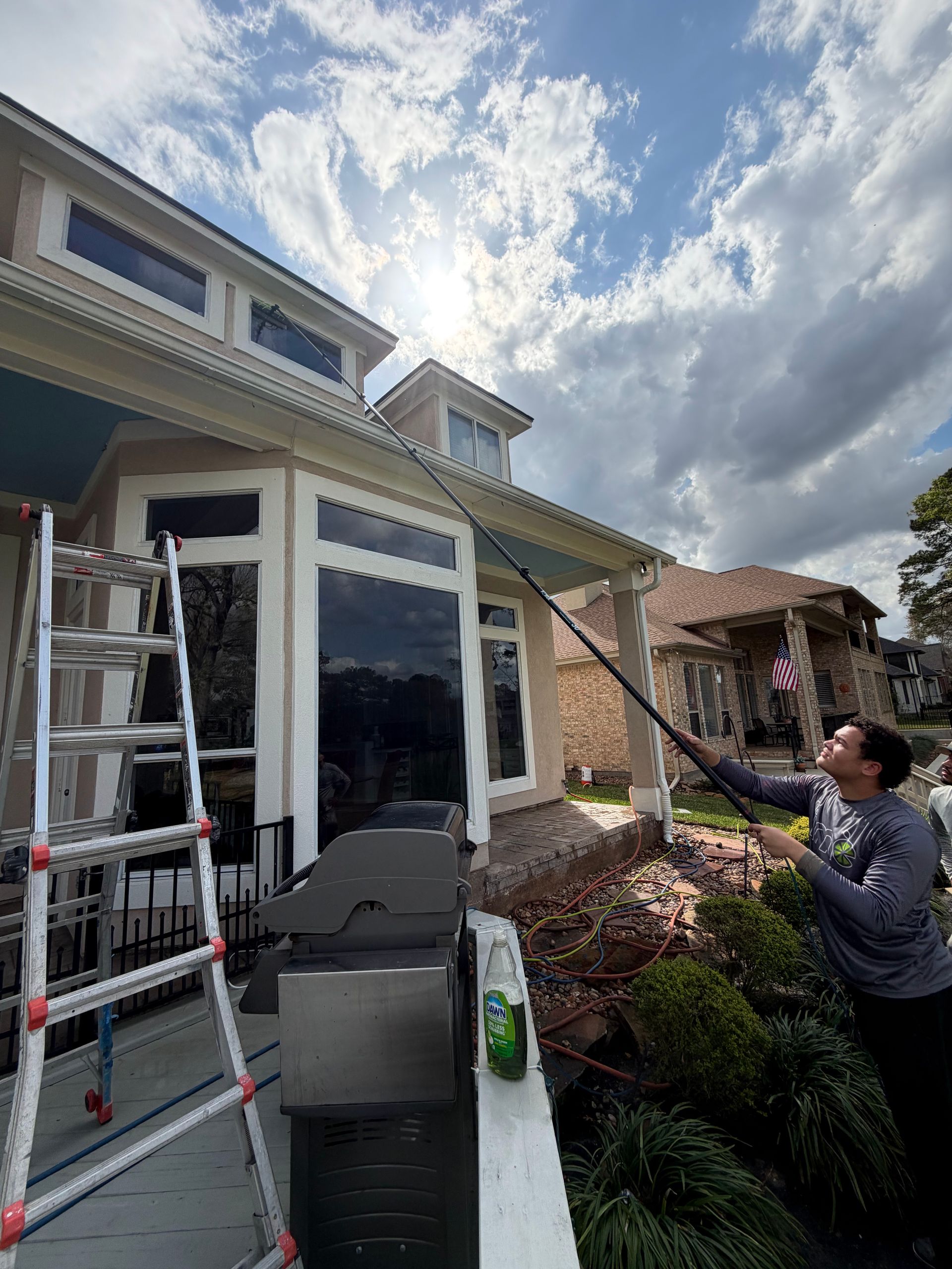 Person using a long pole to clean house windows beside a ladder and gutter on a cloudy day