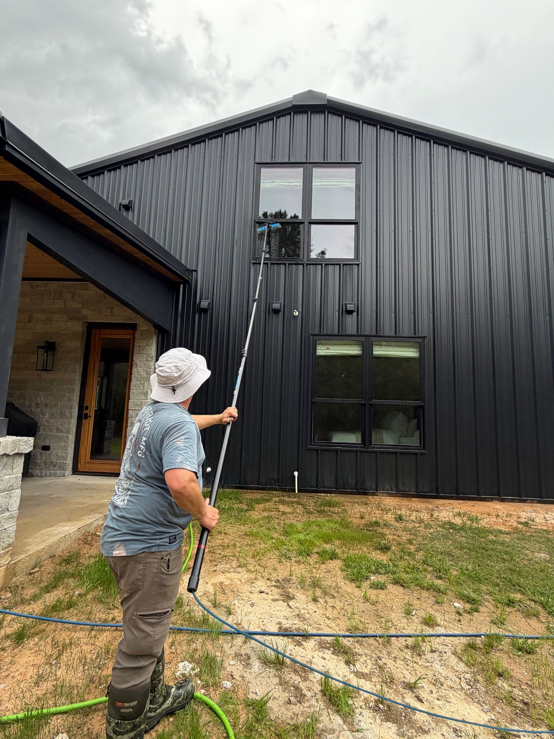 Person using a long pole to wash black house siding and upper windows outside a home