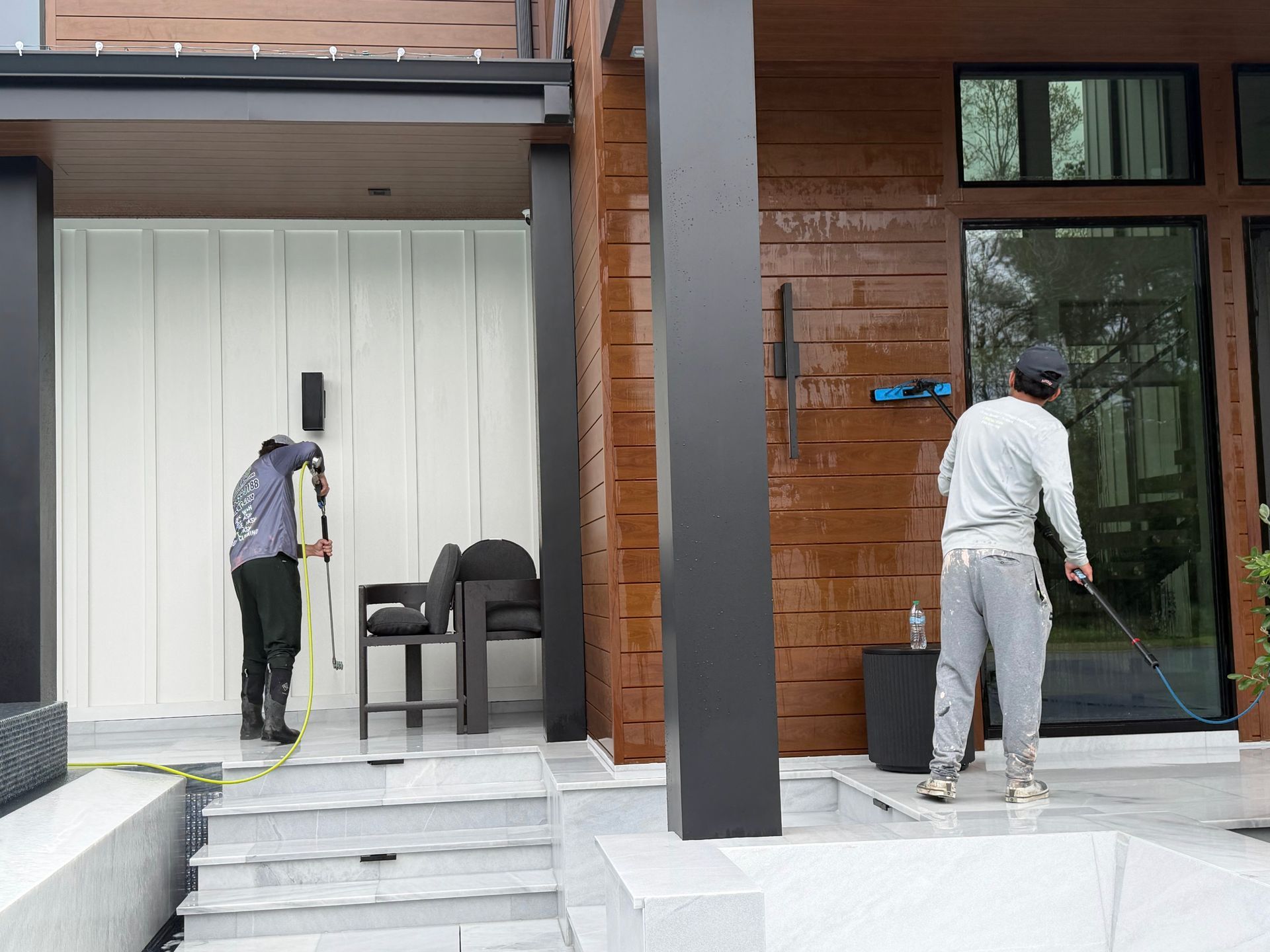 Two workers painting a modern house exterior, one on steps and one by the glass door