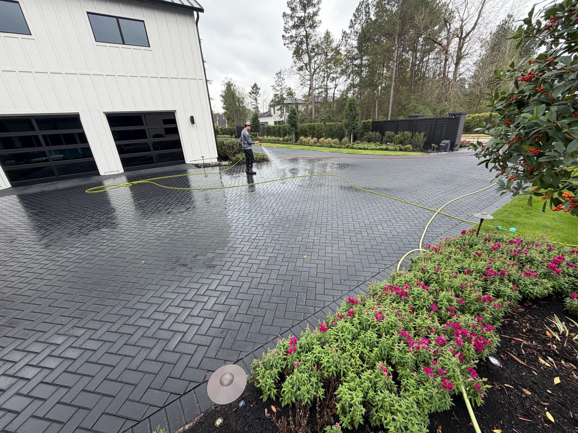 Rainy driveway with a person spraying water near a house and flowerbed。