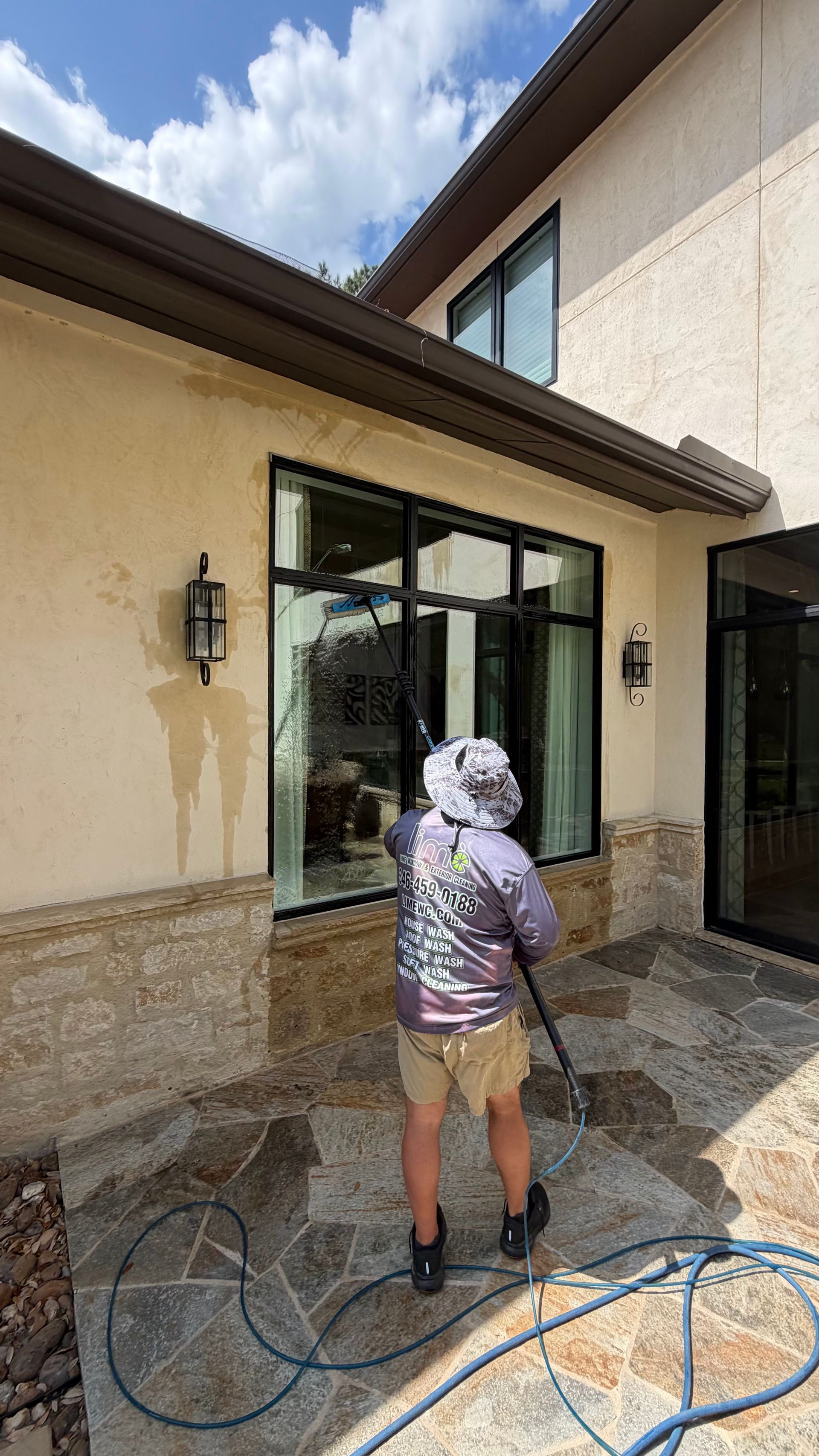 Person pressure-washing windows and patio beside a beige stucco house.
