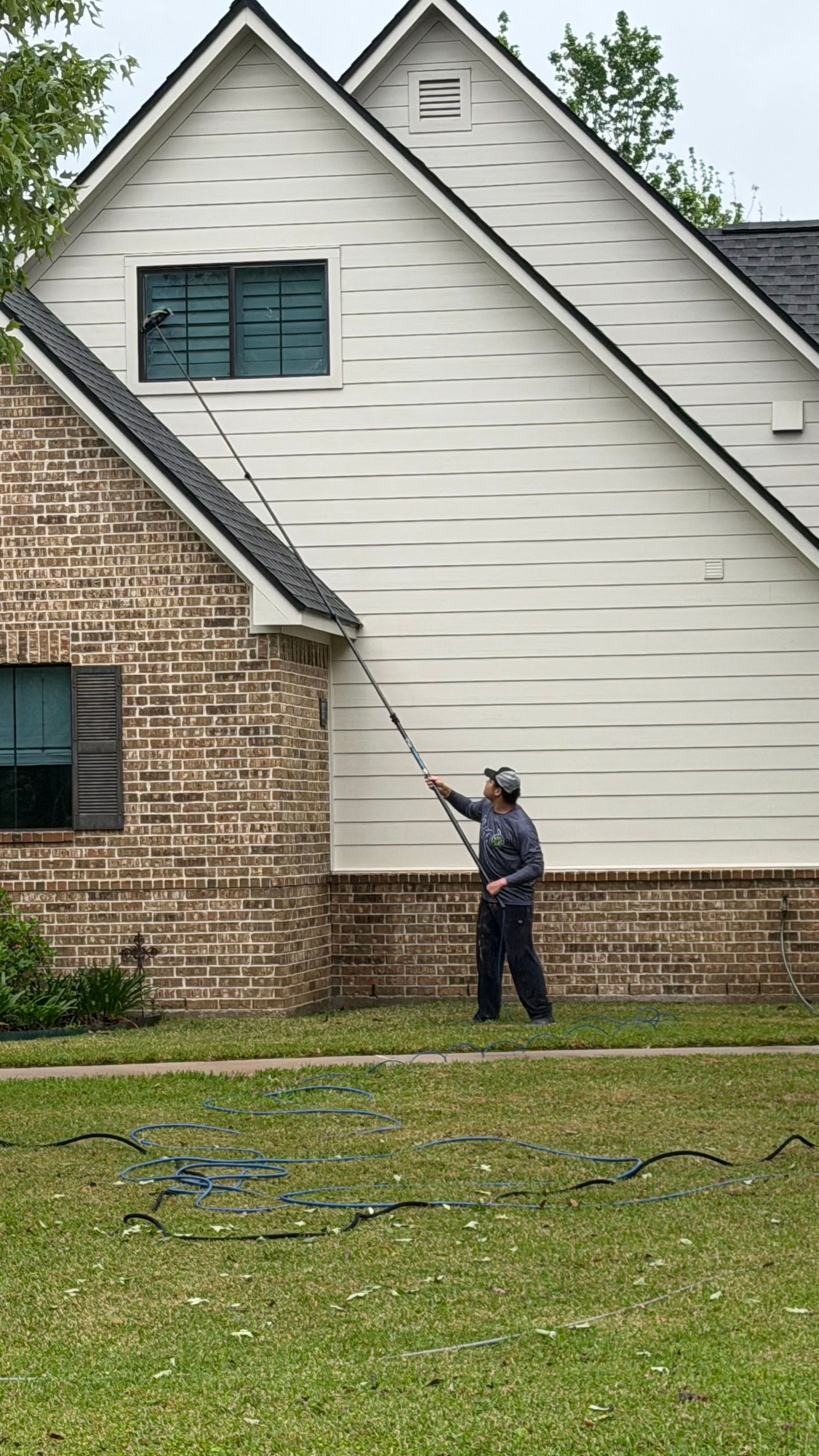 Person pressure-washing a white house wall beside a brick section and green lawn.