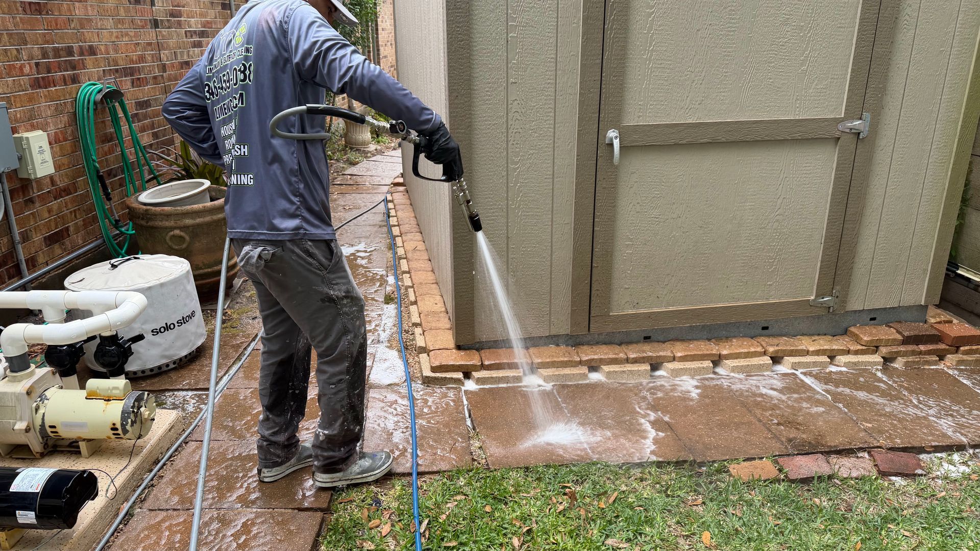 Worker pressure-washing a patio beside a shed, spraying water over brick and grass edge