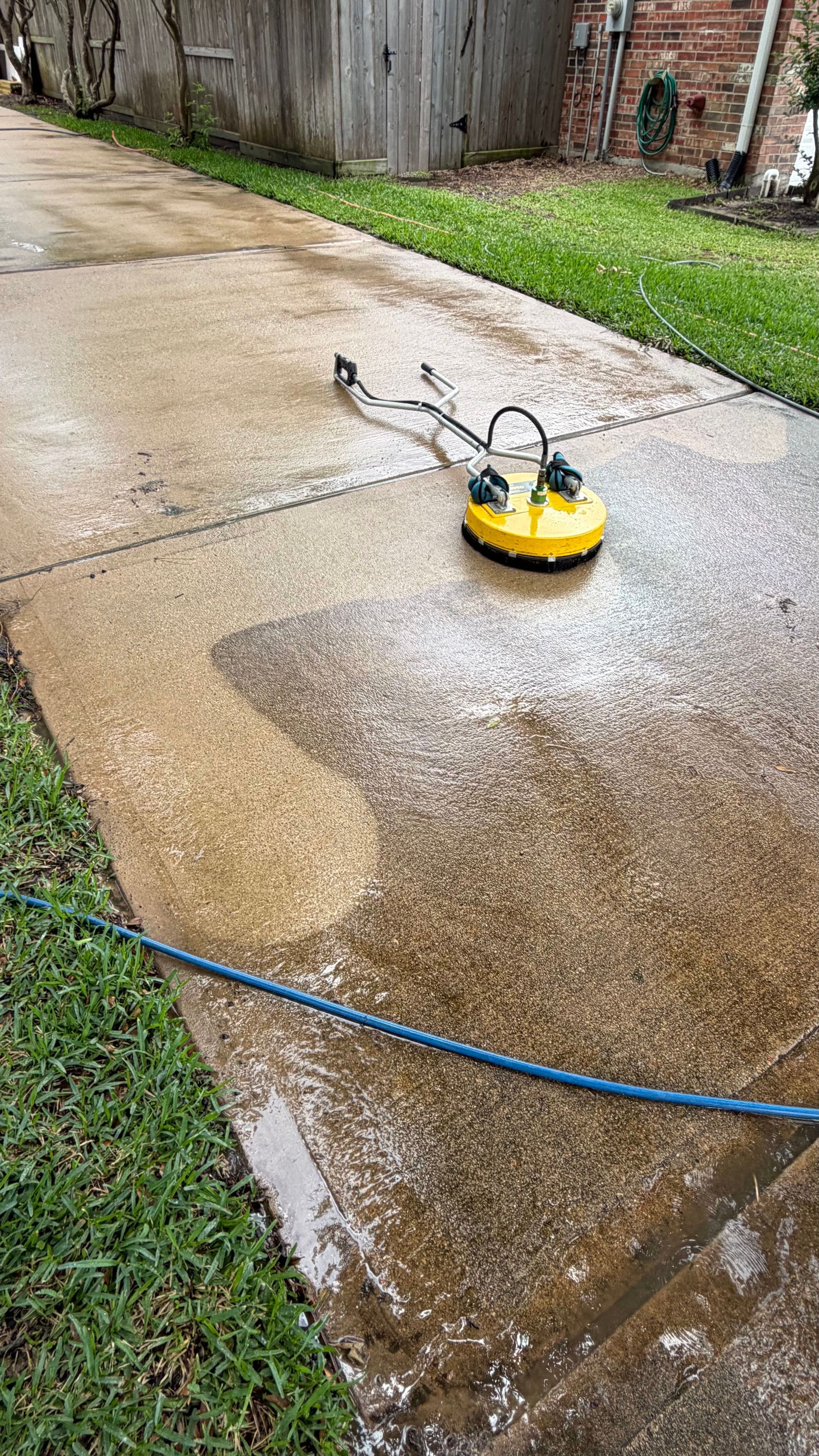 Yellow pressure washer on a wet concrete driveway spraying water near a blue hose line