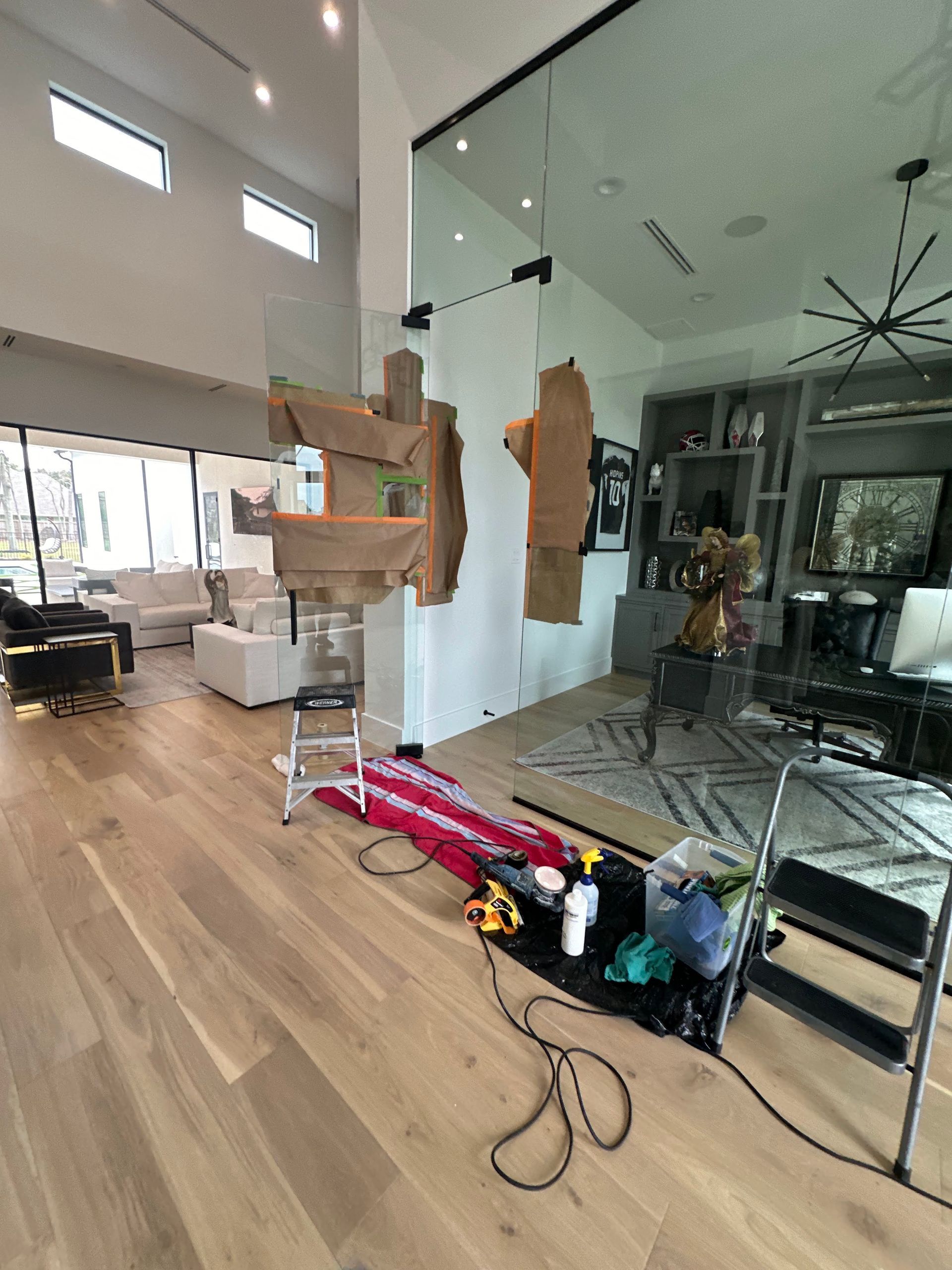 Living room under renovation with glass wall, tools, cables, and a stepladder on hardwood floors