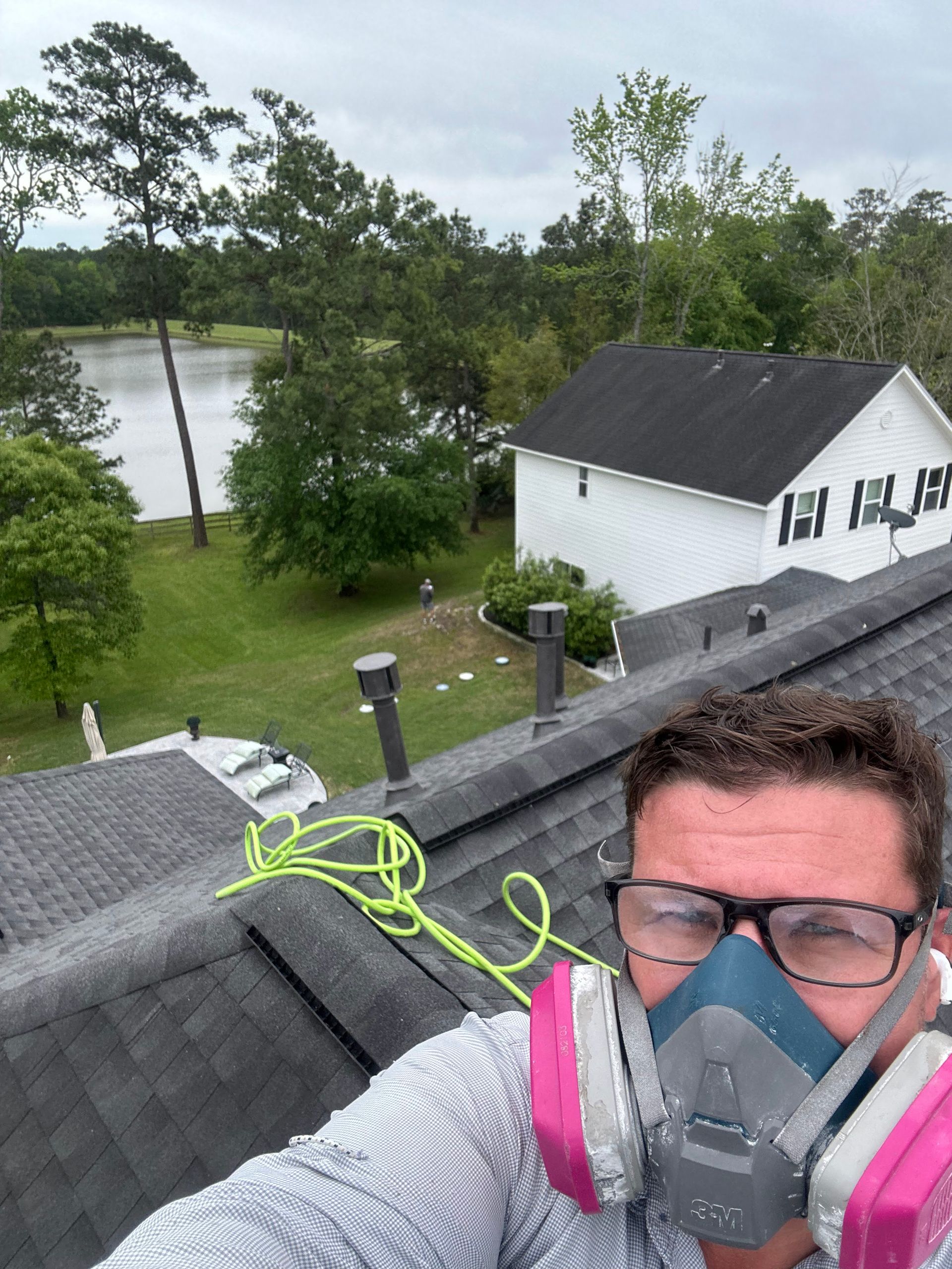 Man wearing respirator and goggles on a rooftop, with trees, a lake, and a white house below.