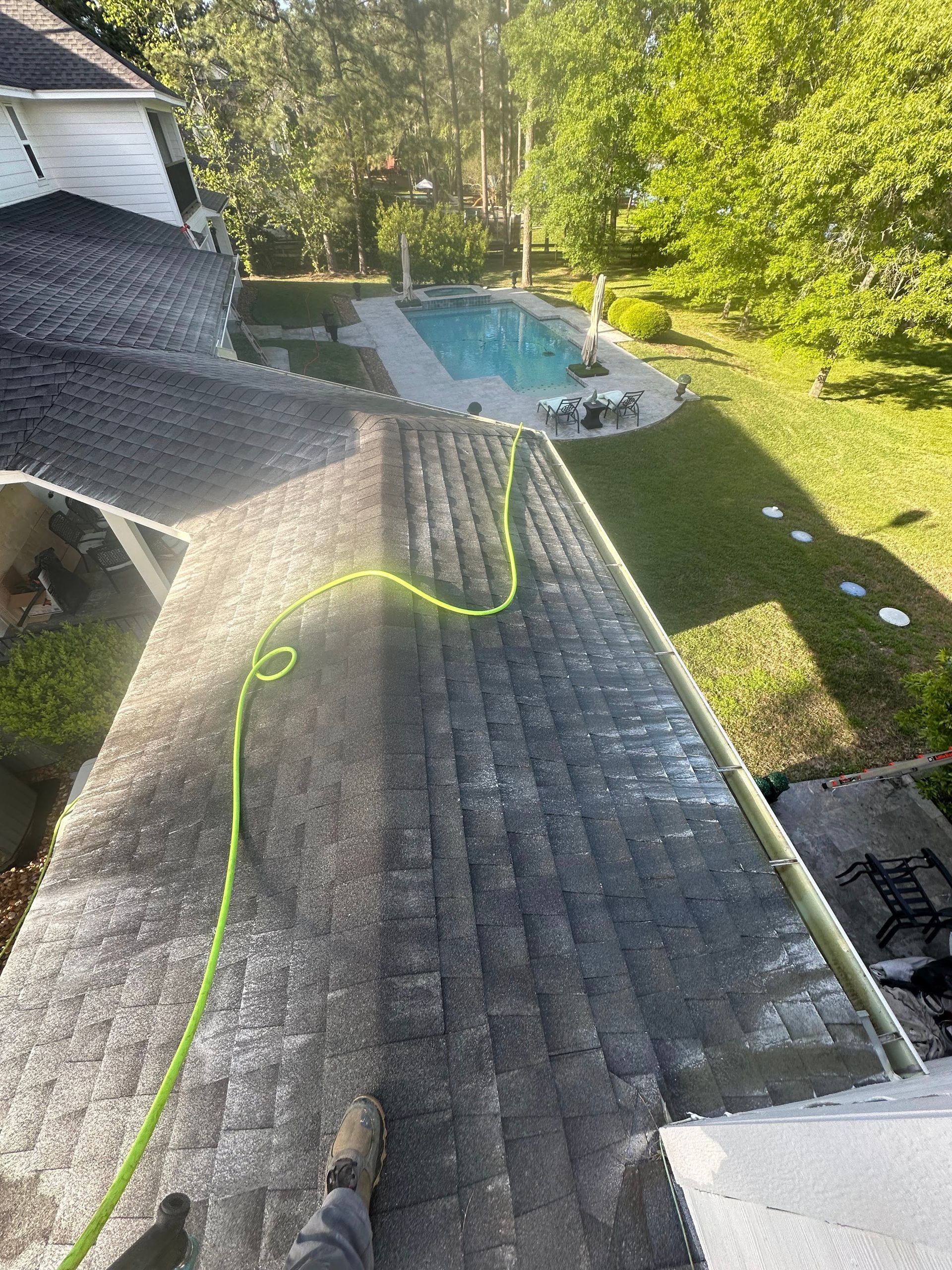 Person washing a gray shingled roof with a hose beside a backyard pool and trees