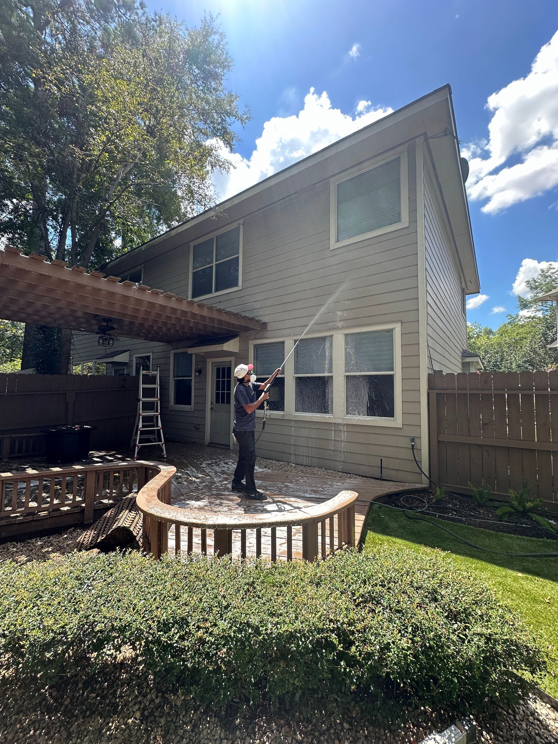 Person standing on a raised brick patio in a sunny backyard beside a two-story house.
