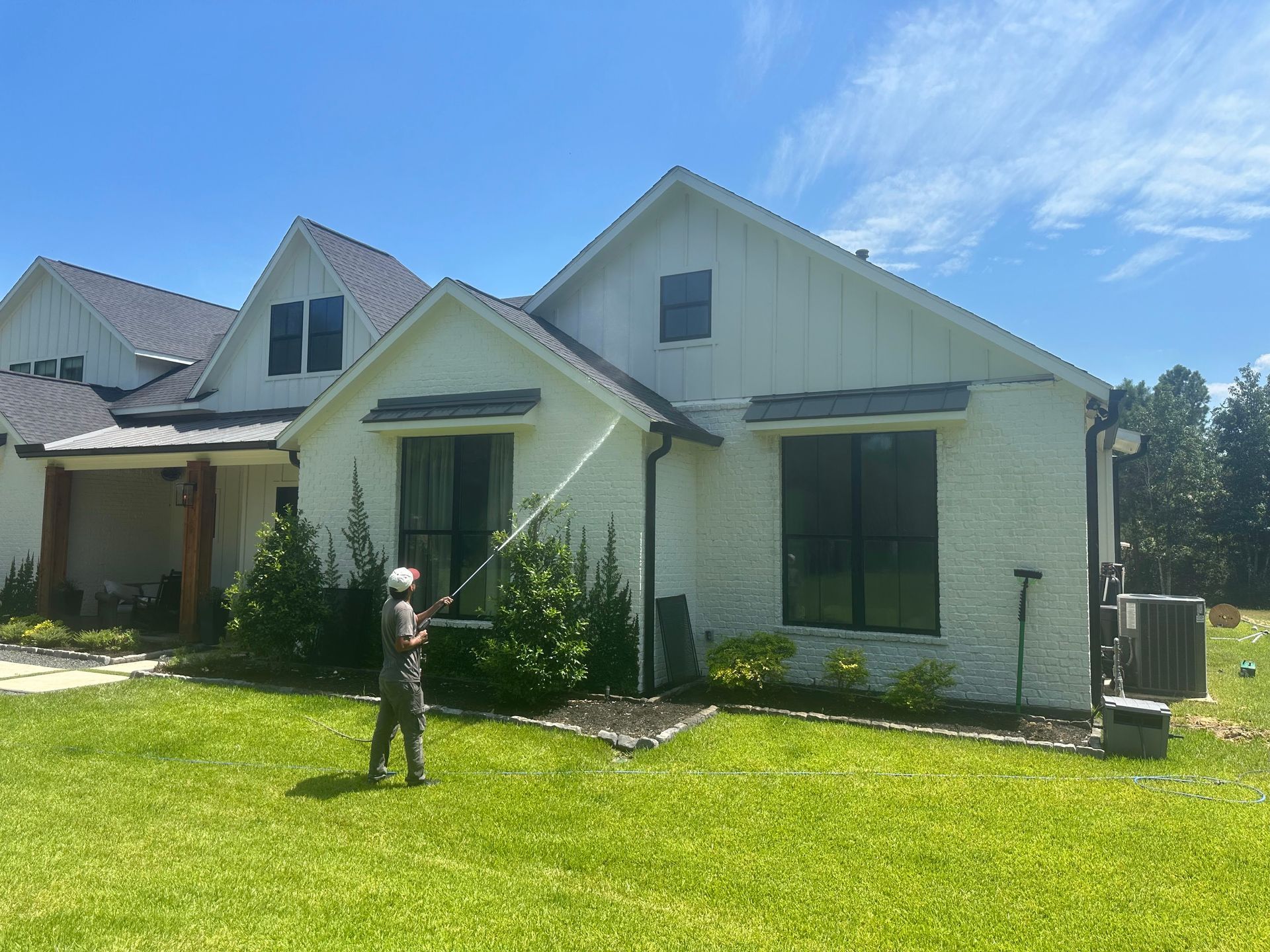Person watering plants in front of a light-colored suburban house with a green lawn.