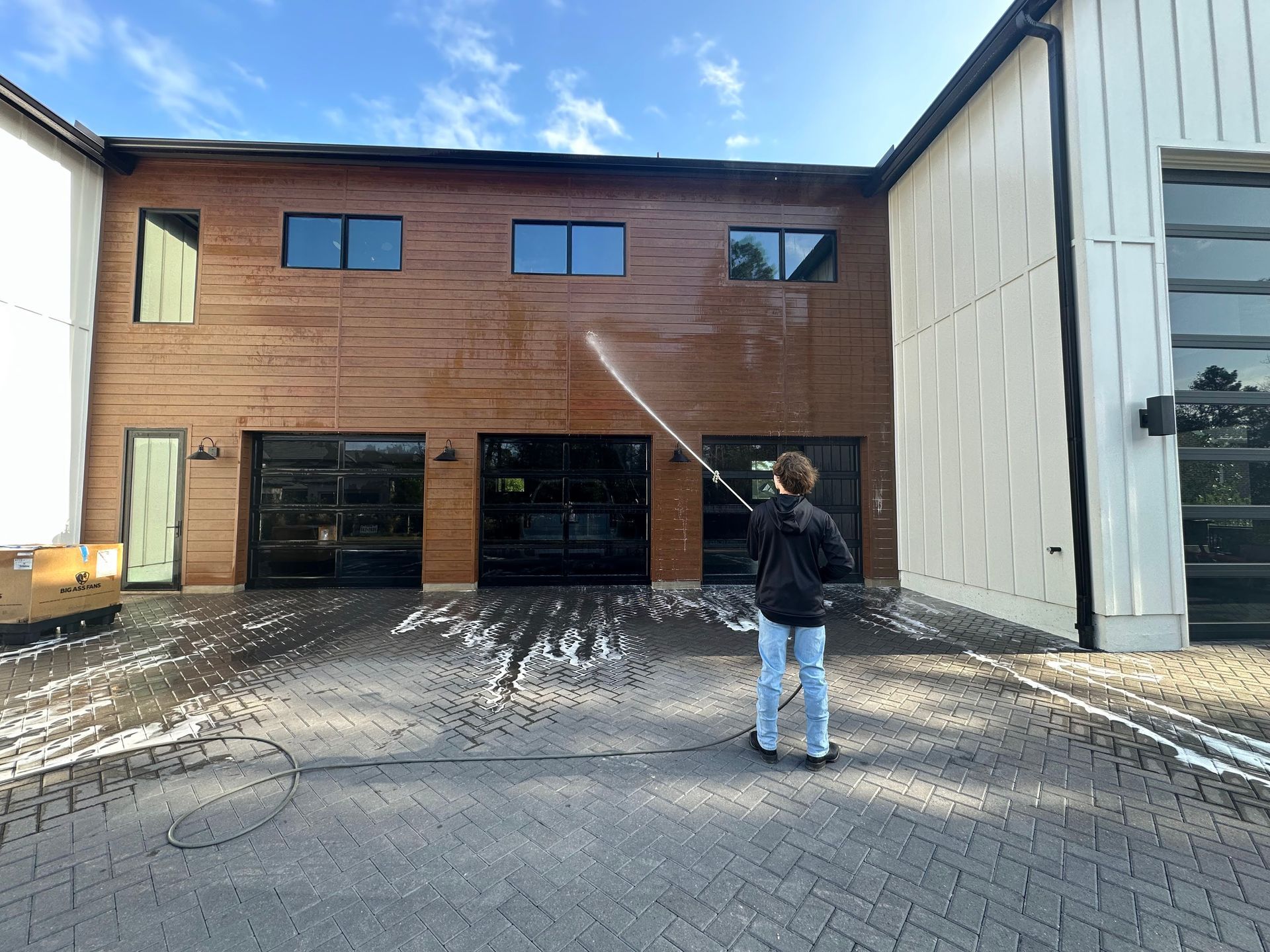 Person spraying water on a two-story wooden building with black garage doors and large windows.