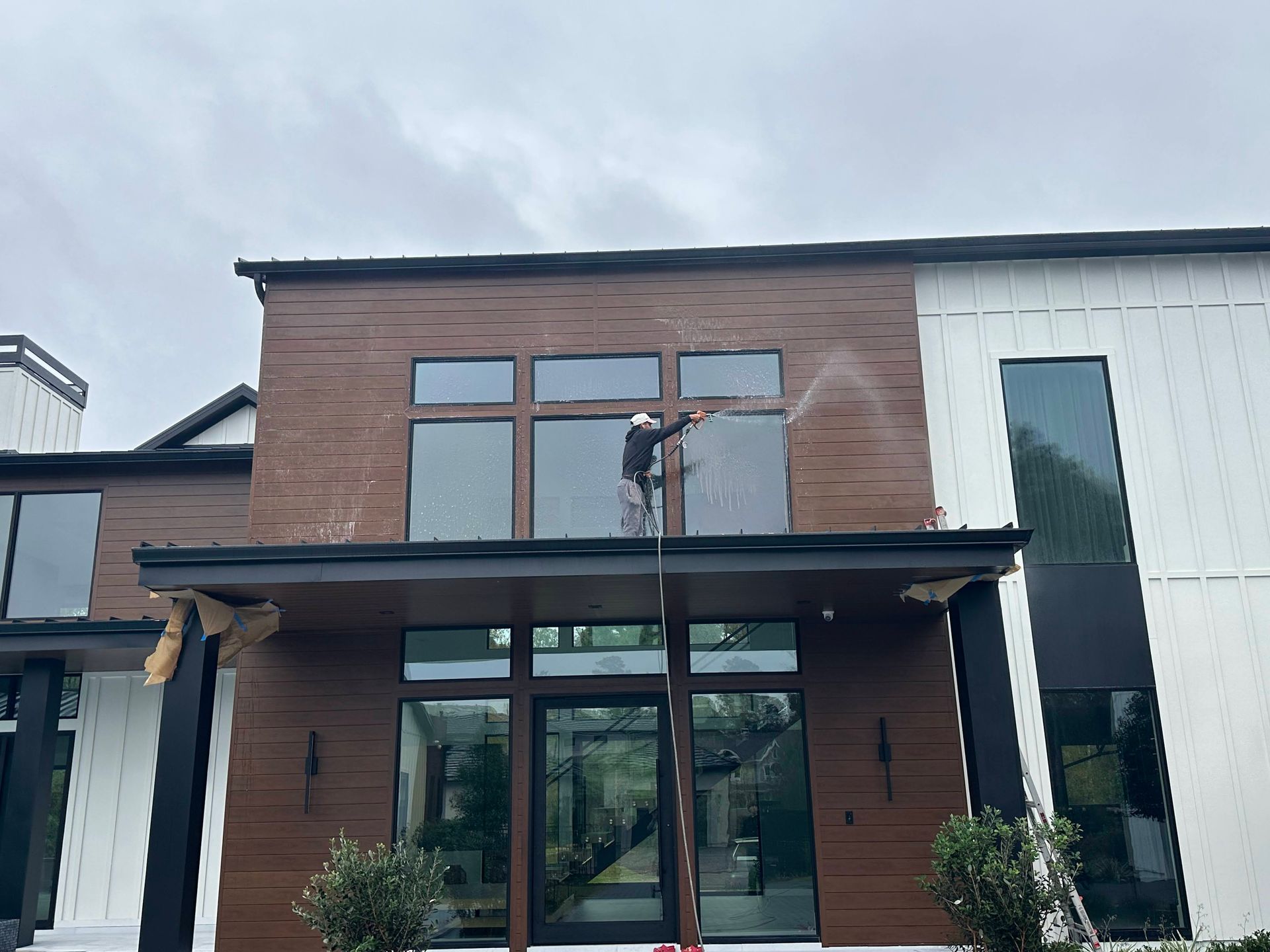 Modern two-story house with wood siding and large windows; worker on upper balcony installing something