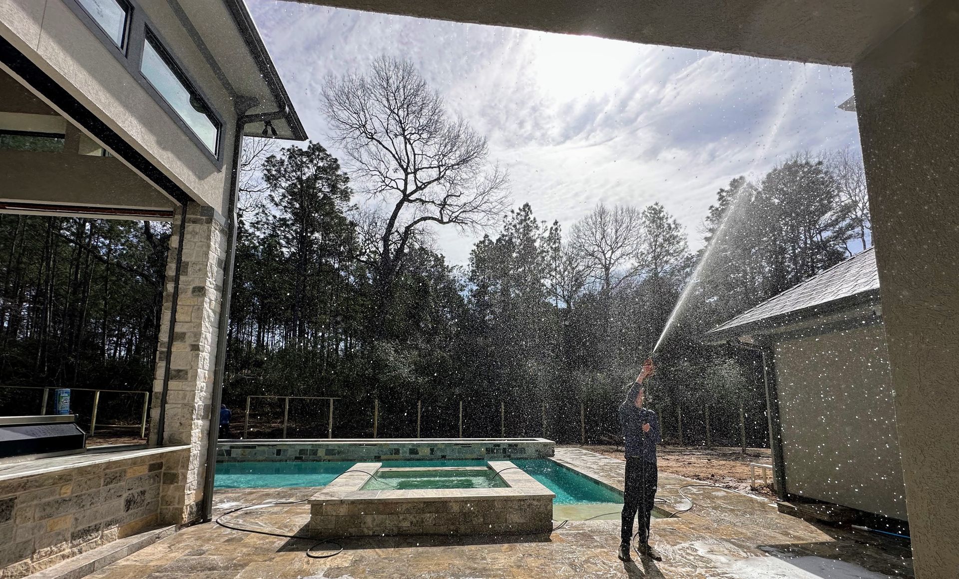 Backyard pool with a person spraying water from a hose beside a house and trees