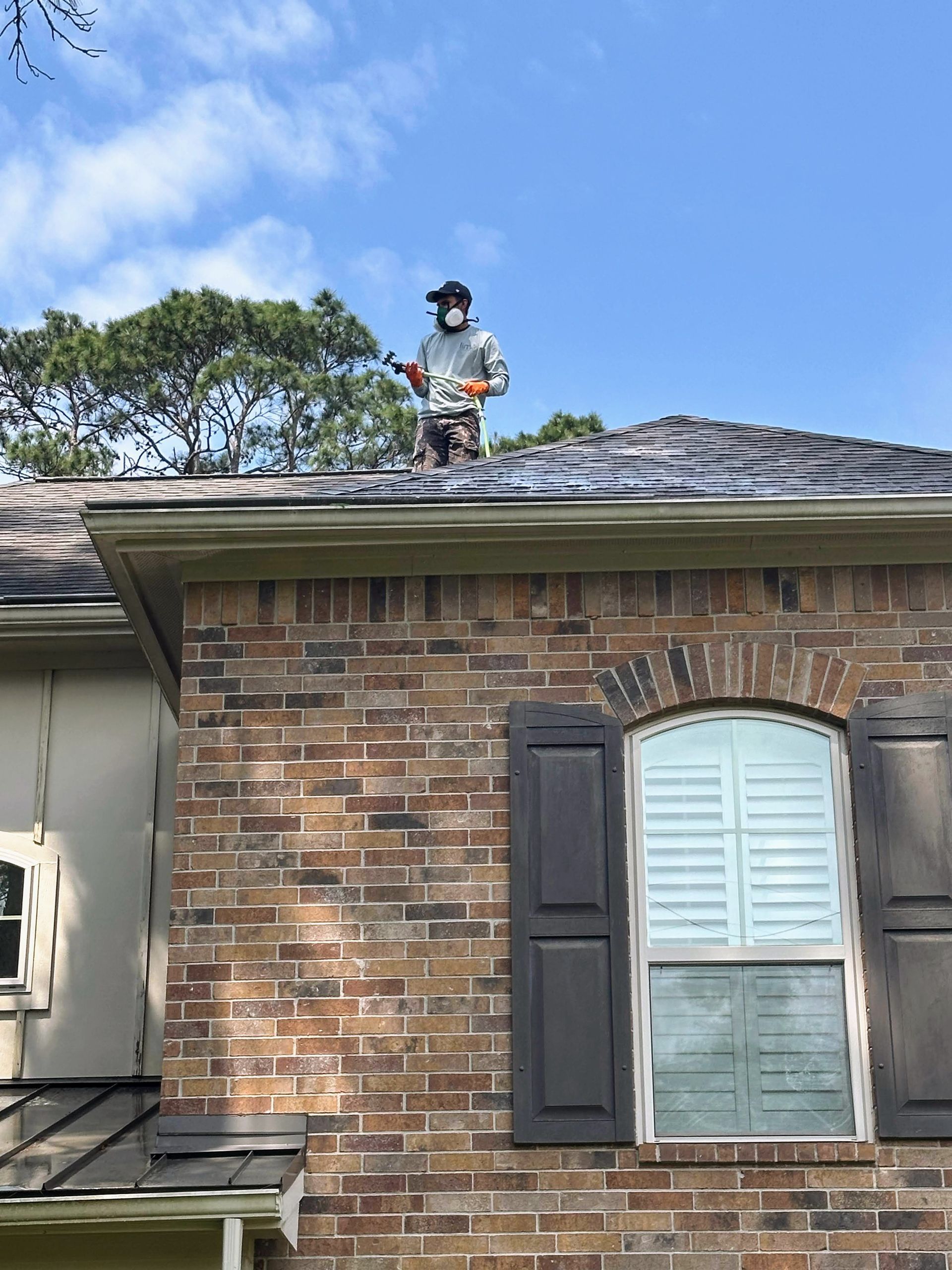 Person on a roof inspecting shingles on a brick house under a clear blue sky