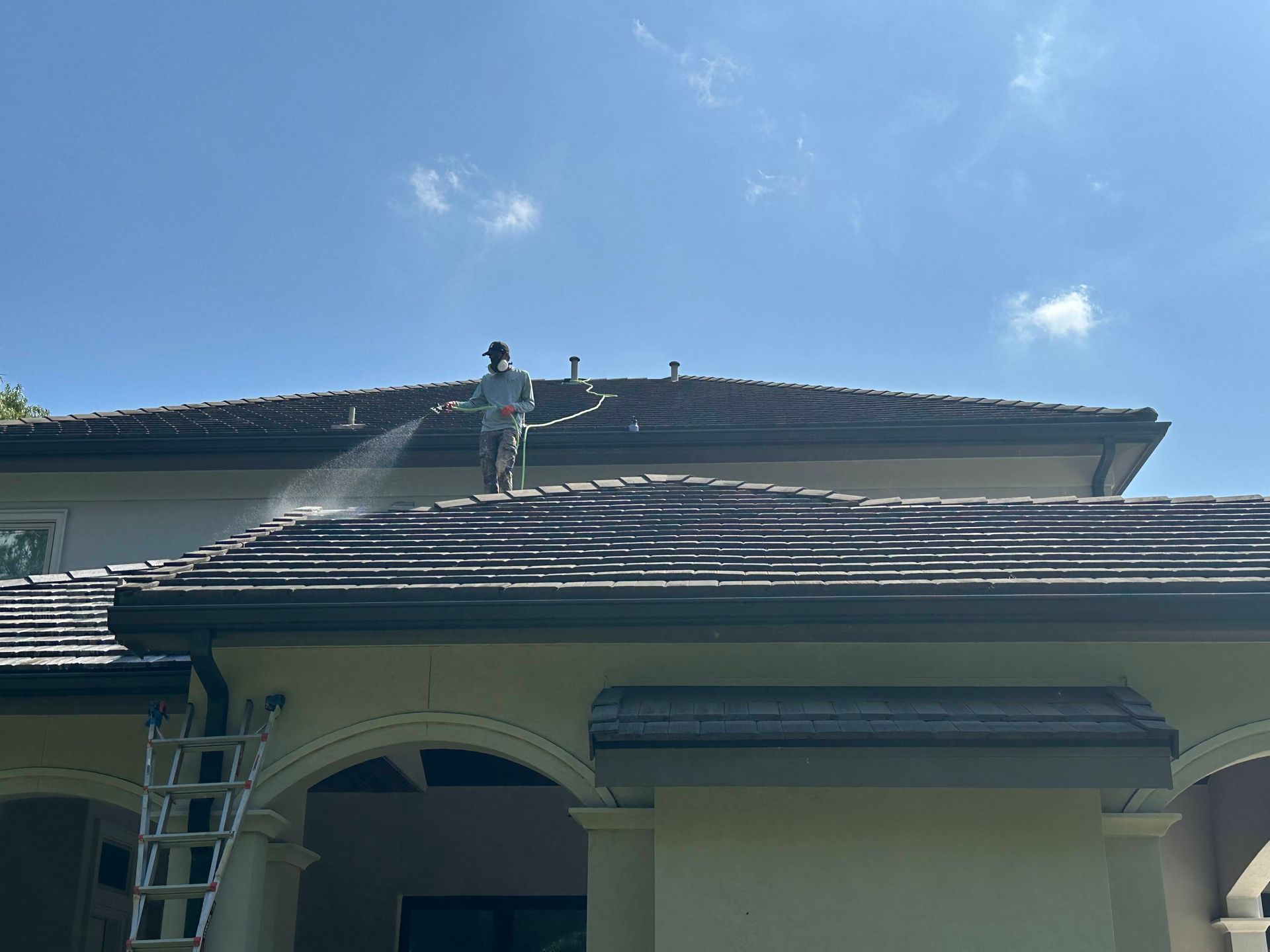 Worker pressure-washing a house roof, with a ladder and bright blue sky above
