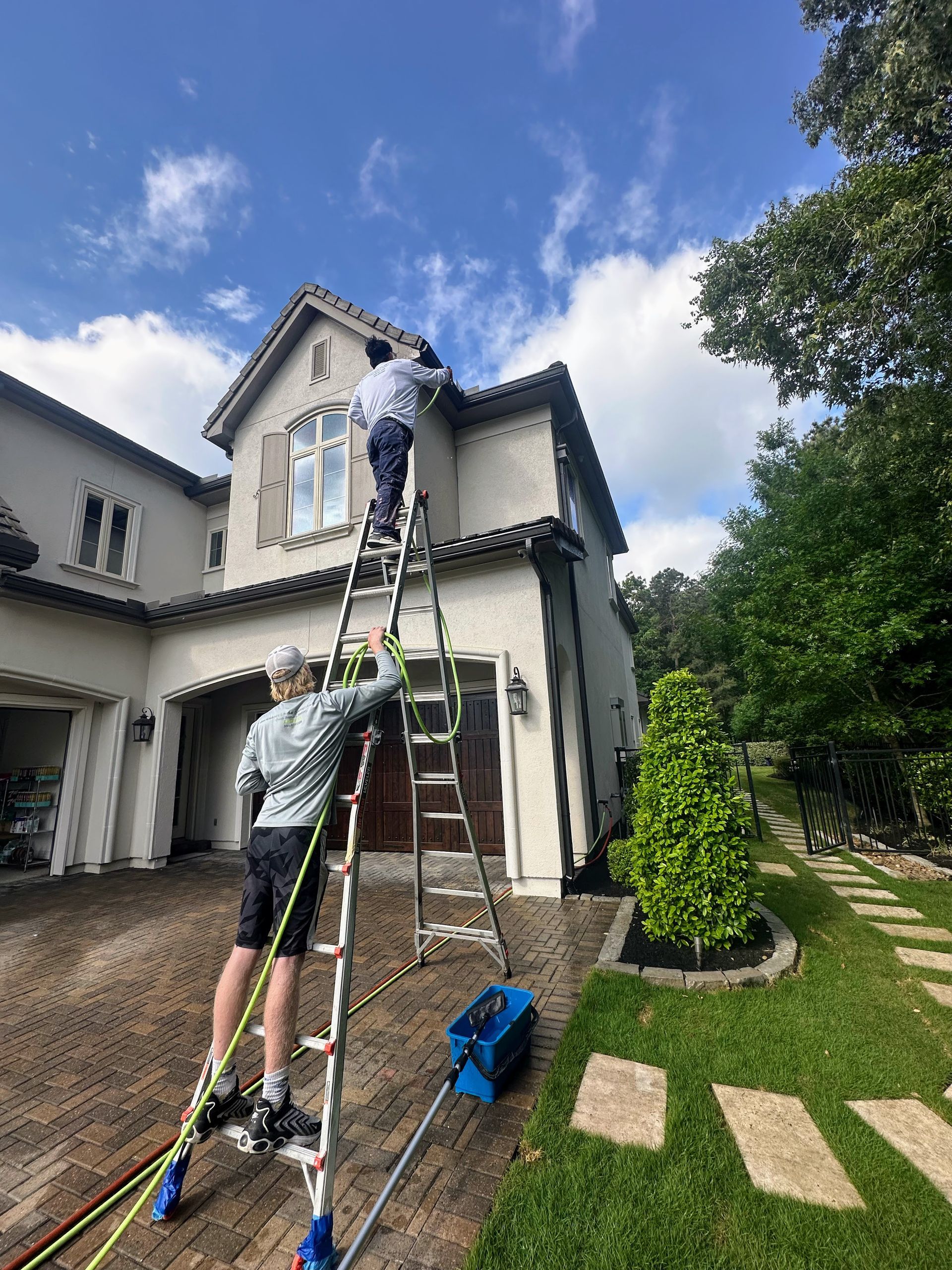 Workers on ladders painting a two-story house exterior under a blue sky.