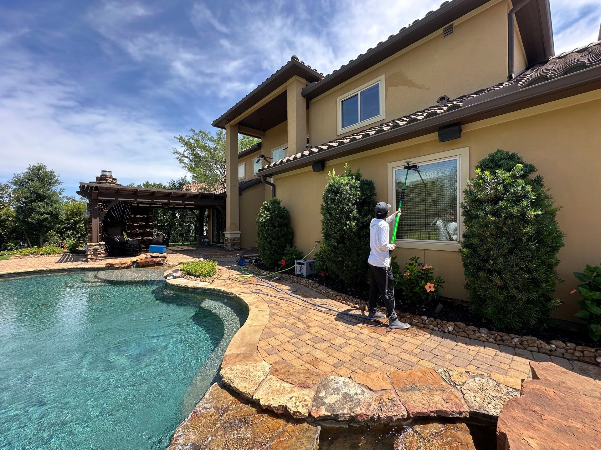 Poolside patio beside a tan house, with a person trimming shrubs near a window.