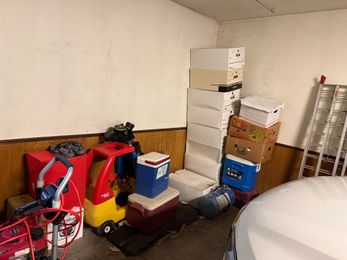 Cluttered garage with stacked boxes, a red ride-on toy car, and folded metal chairs against the wall
