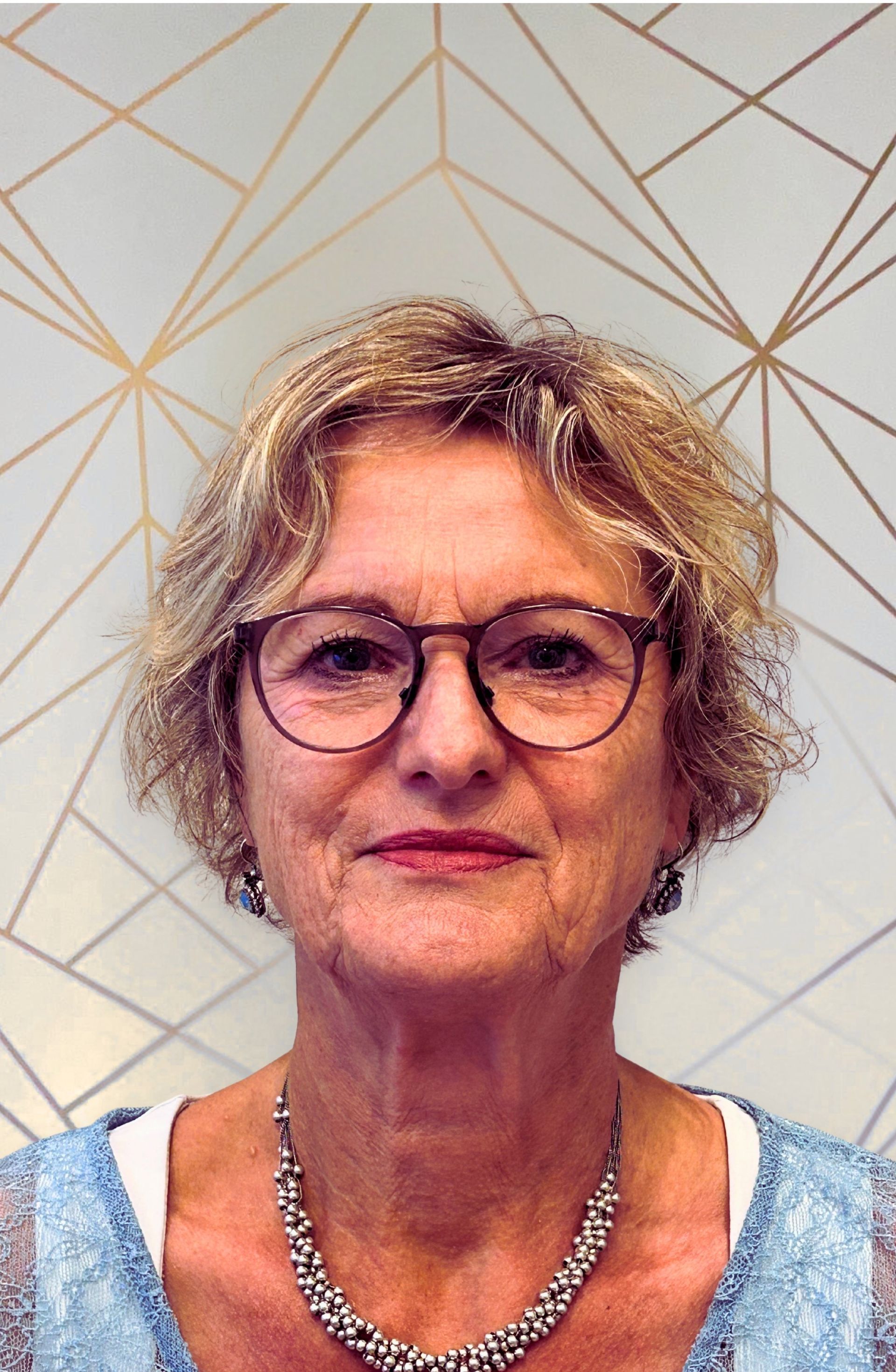 Vrouw met een bril en kort, krullend blond haar, draagt een ketting en een lichtblauwe top.