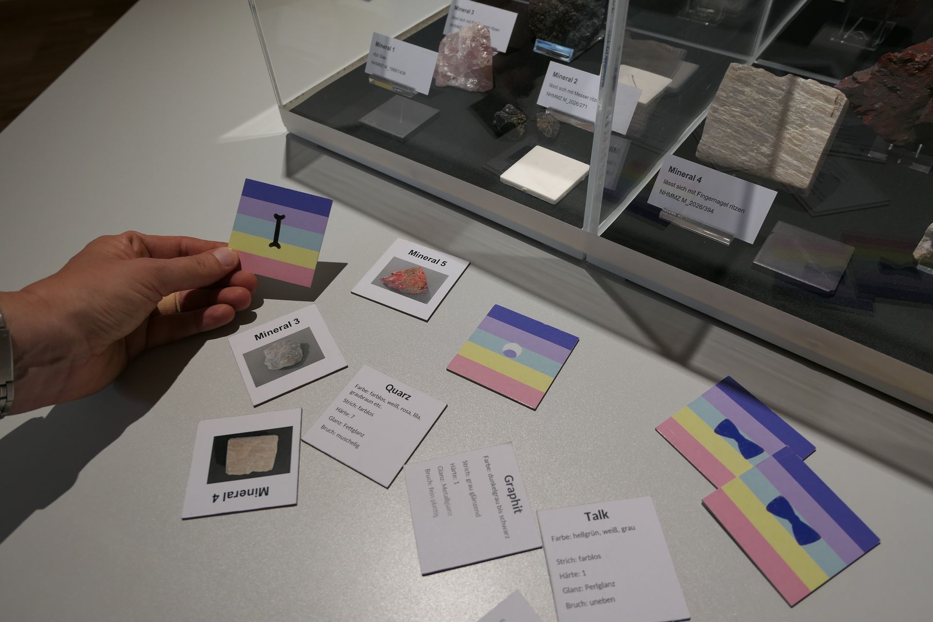 Eine Hand hält eine bunte Karte über einen Tisch mit verschiedenen bedruckten Karten und Papieren vor einer Vitrine.