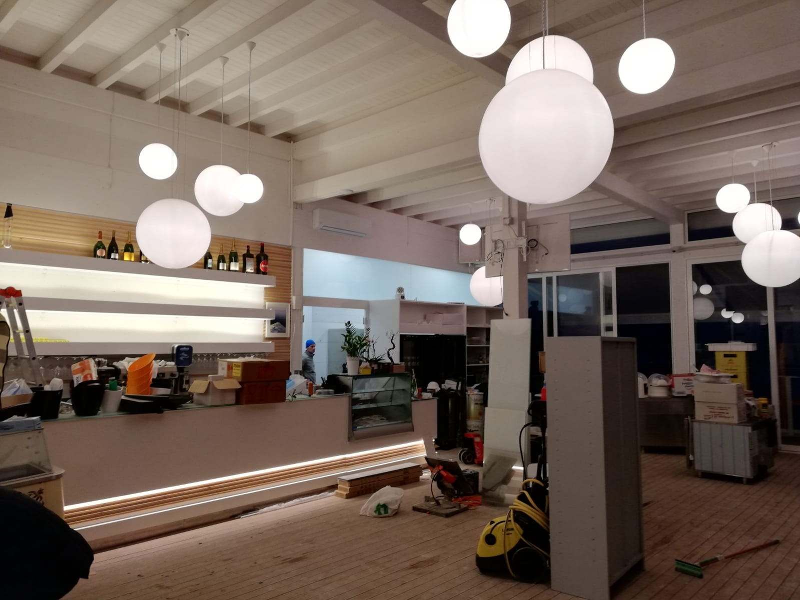 Interno di un bar con grandi lampade a globo bianche. Sono visibili un bancone e alcuni detriti edili.