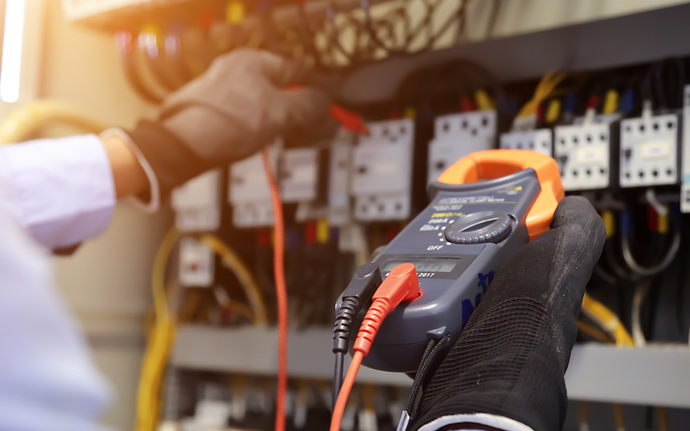Elettricista che utilizza un multimetro per controllare i cavi in un quadro elettrico, indossando guanti da lavoro.