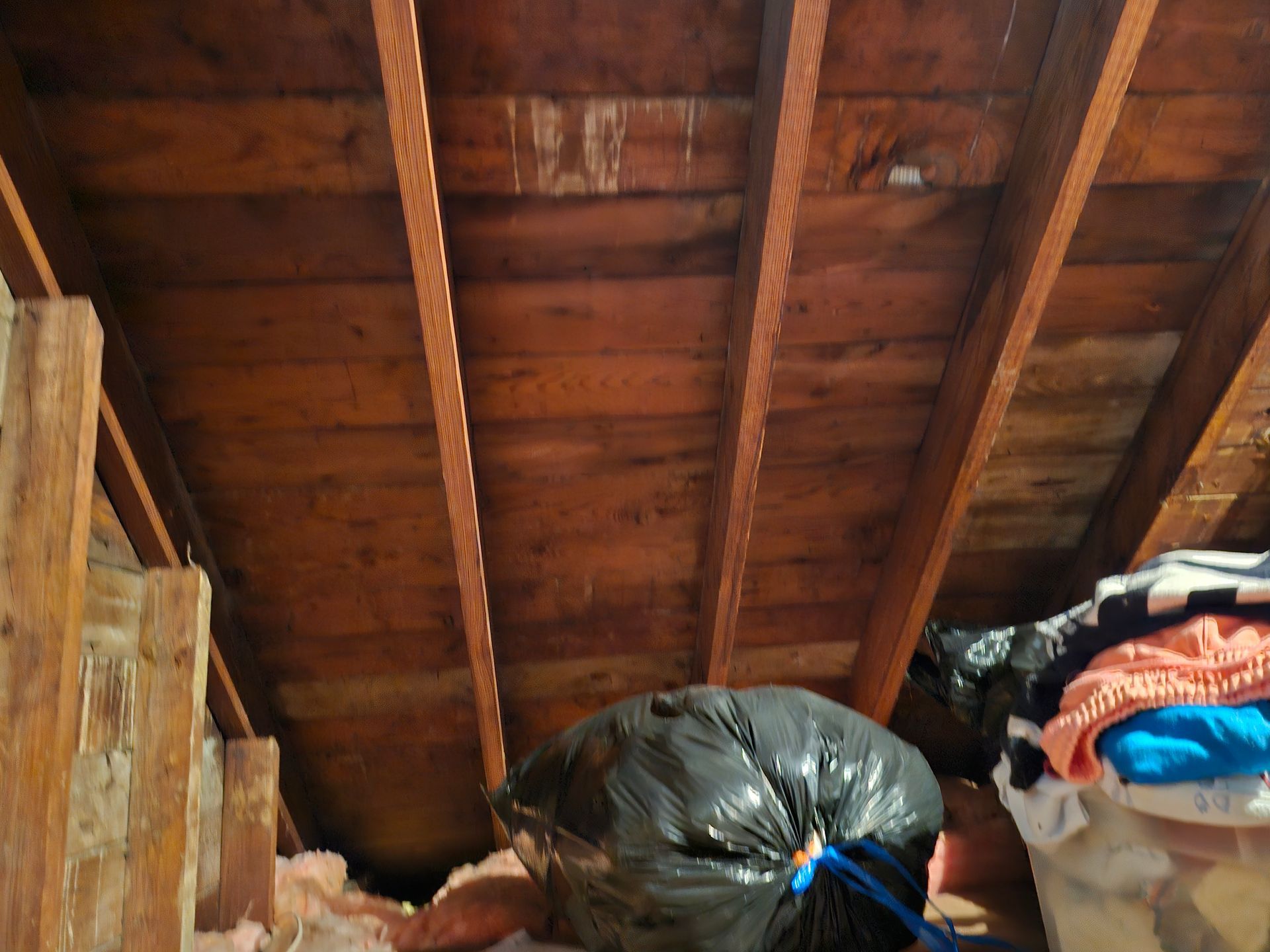 Wooden attic ceiling with exposed beams and a black trash bag amid stored items.