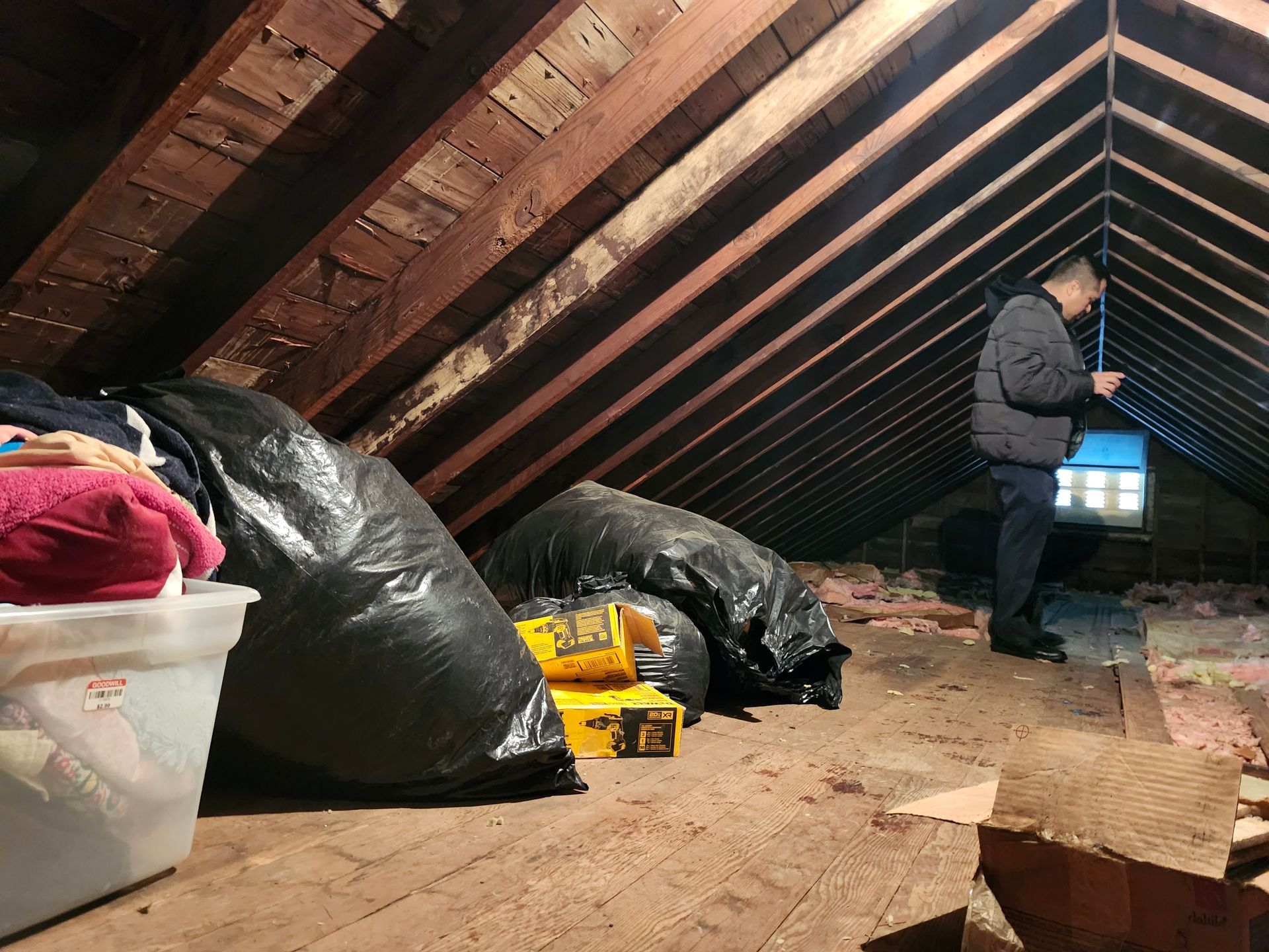 Person in an attic beside black trash bags, storage bins, and a yellow tool case under slanted rafters.
