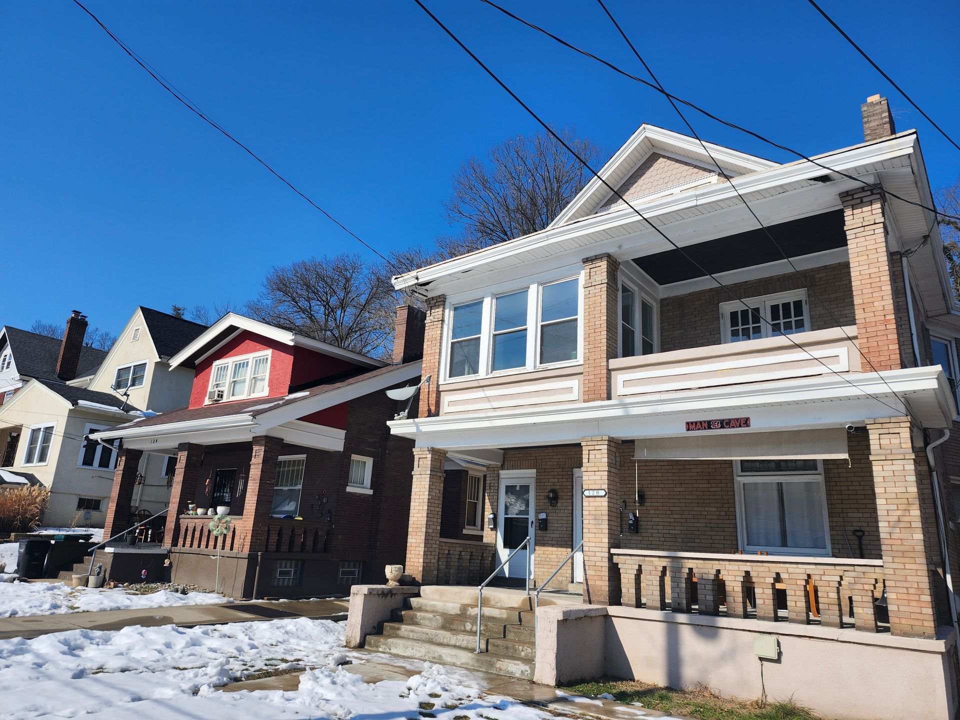 Snow-covered houses on a sunny street, including a white and brick home with a porch.