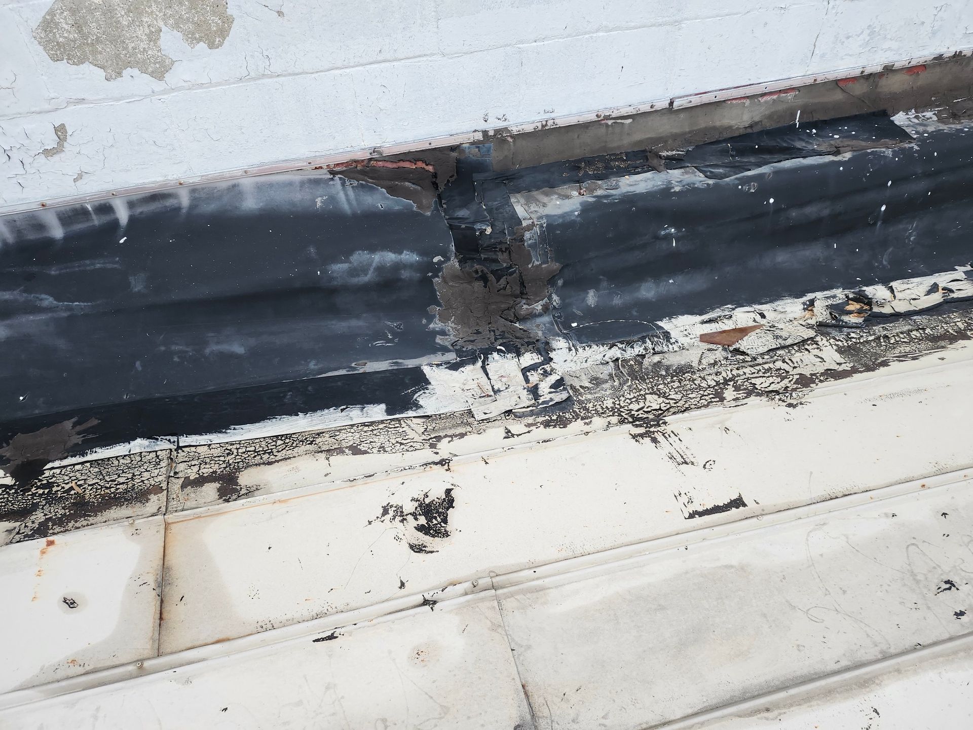 Damaged black roof flashing with cracked sealant above white siding, showing peeling and debris