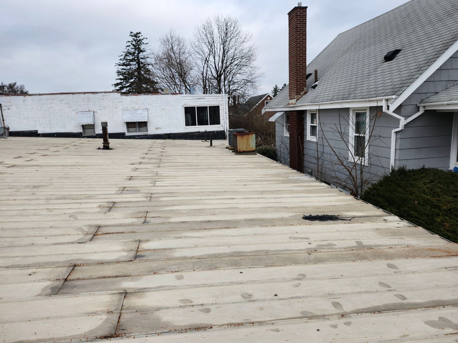 Snow-covered flat rooftop beside a gray house and brick chimney on an overcast day