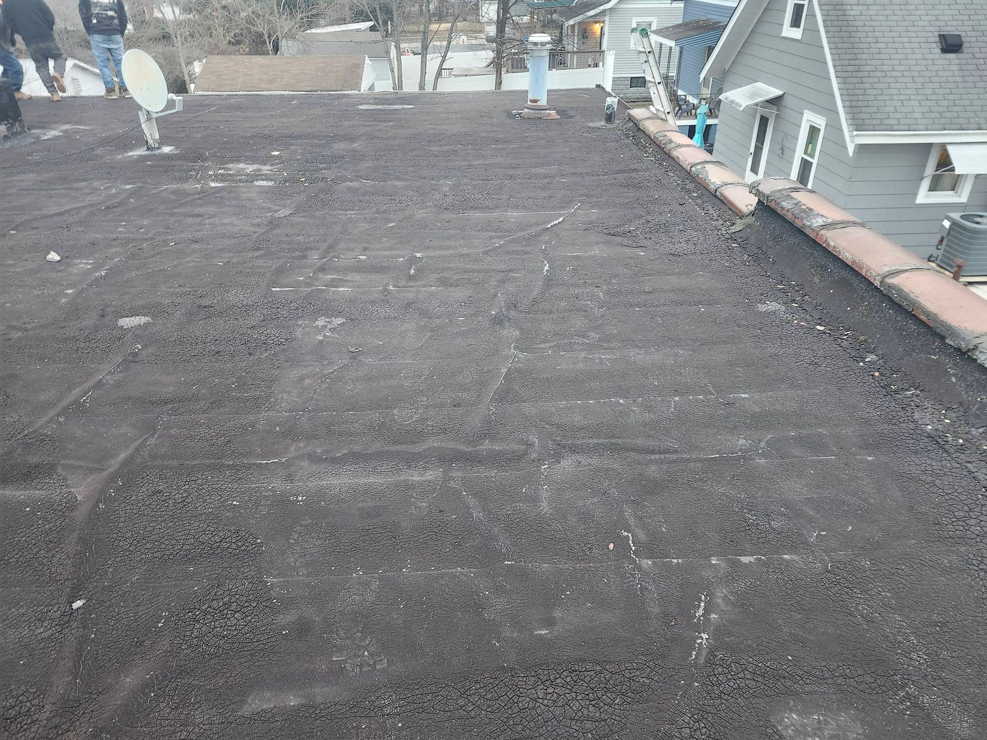 Flat dark roof under construction with workers and a neighboring house visible in the background.