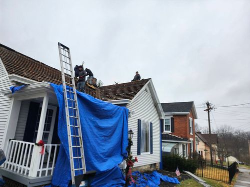 Workers on a roof repairing storm damage to a white house draped with a blue tarp