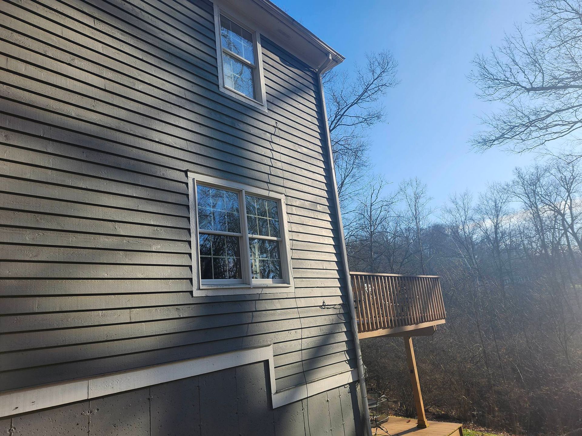 Side of a beige house with windows and a small wooden deck overlooking a wooded area