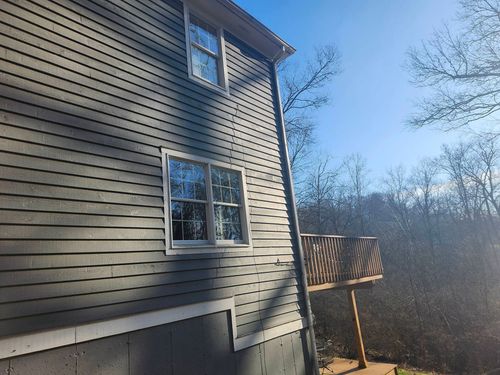 Side of a beige house with windows and a small wooden deck, beside leafless trees under a blue sky