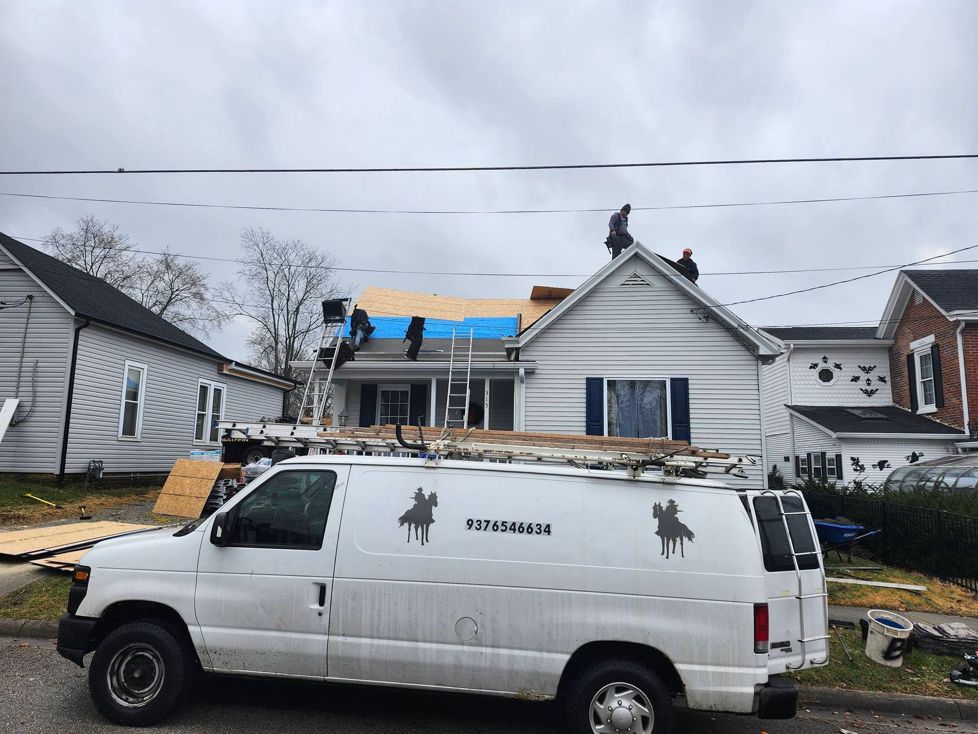 White plumbing van parked in front of a house with roof repair work underway under a cloudy sky