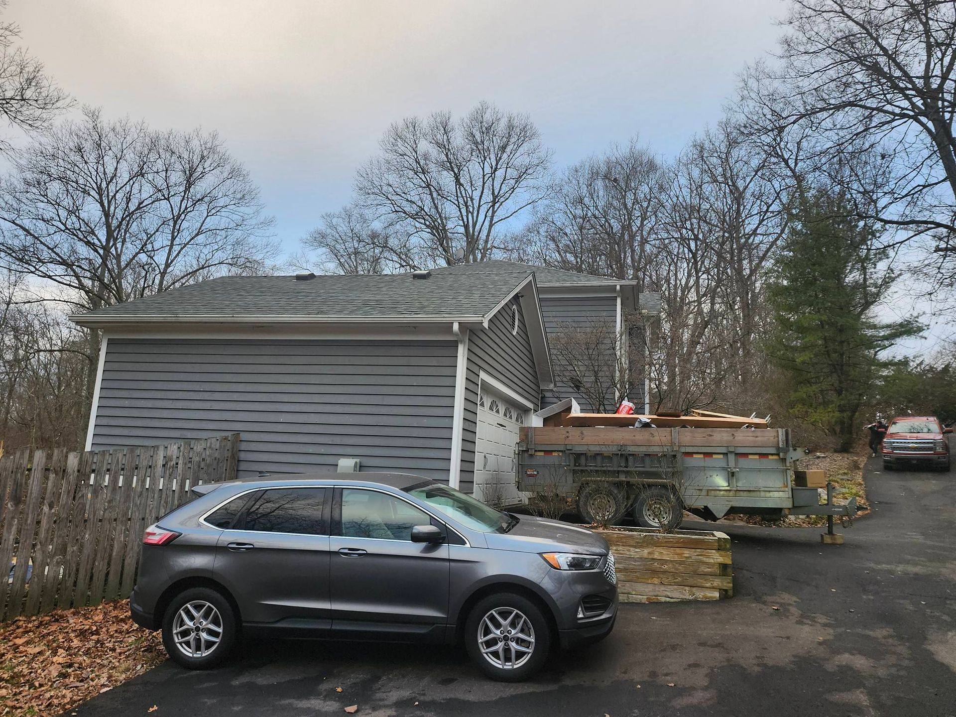 Gray SUV parked by a gray house with a trailer and woodpile in a driveway under bare trees