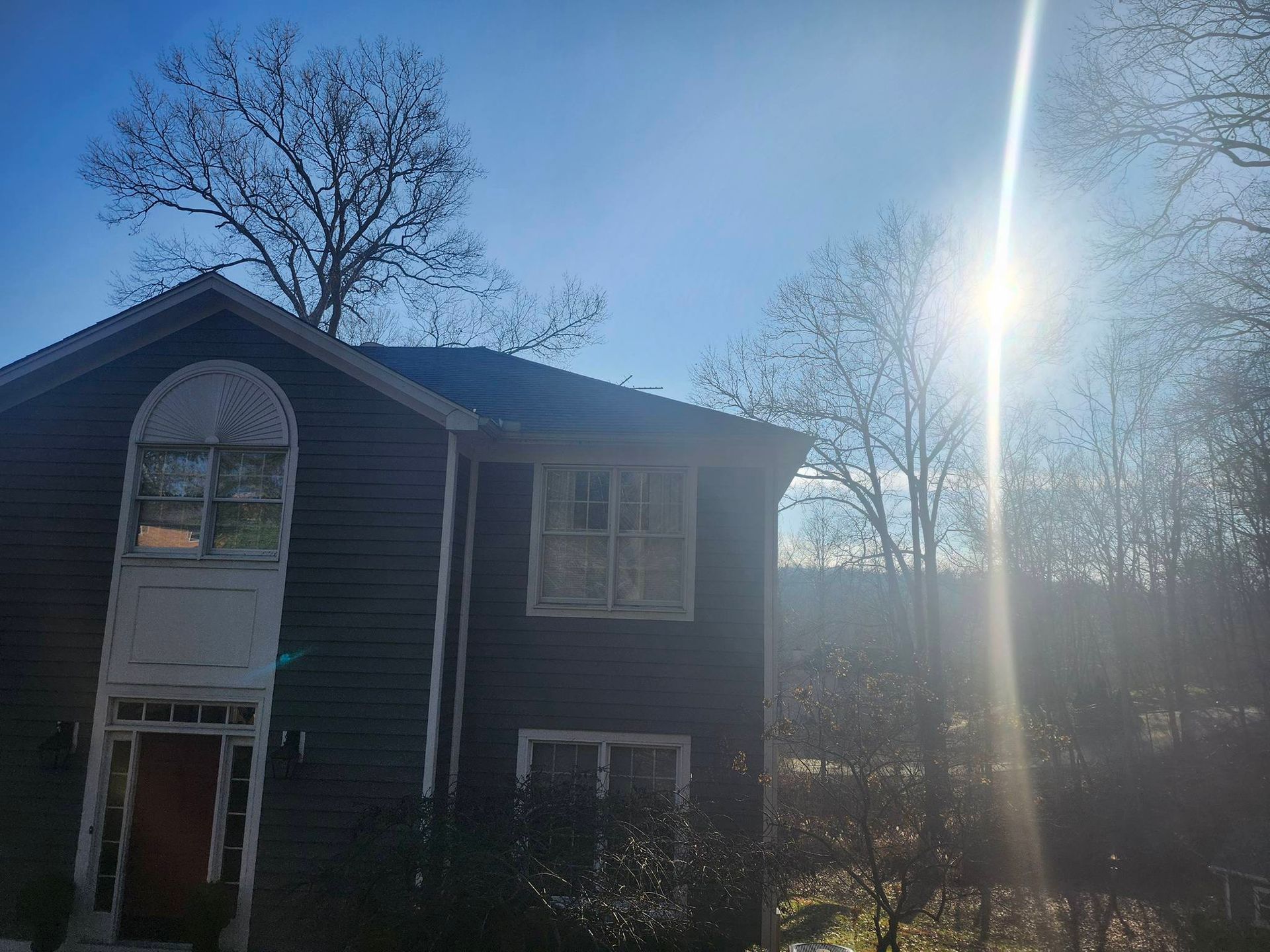 Dark two-story house with bare trees at sunset, bright sun glare on the right