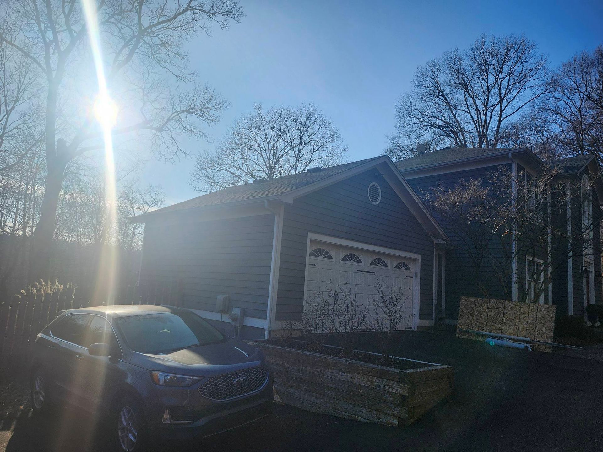 Blue SUV parked in a sunlit driveway beside a brick house with garage door and trees.