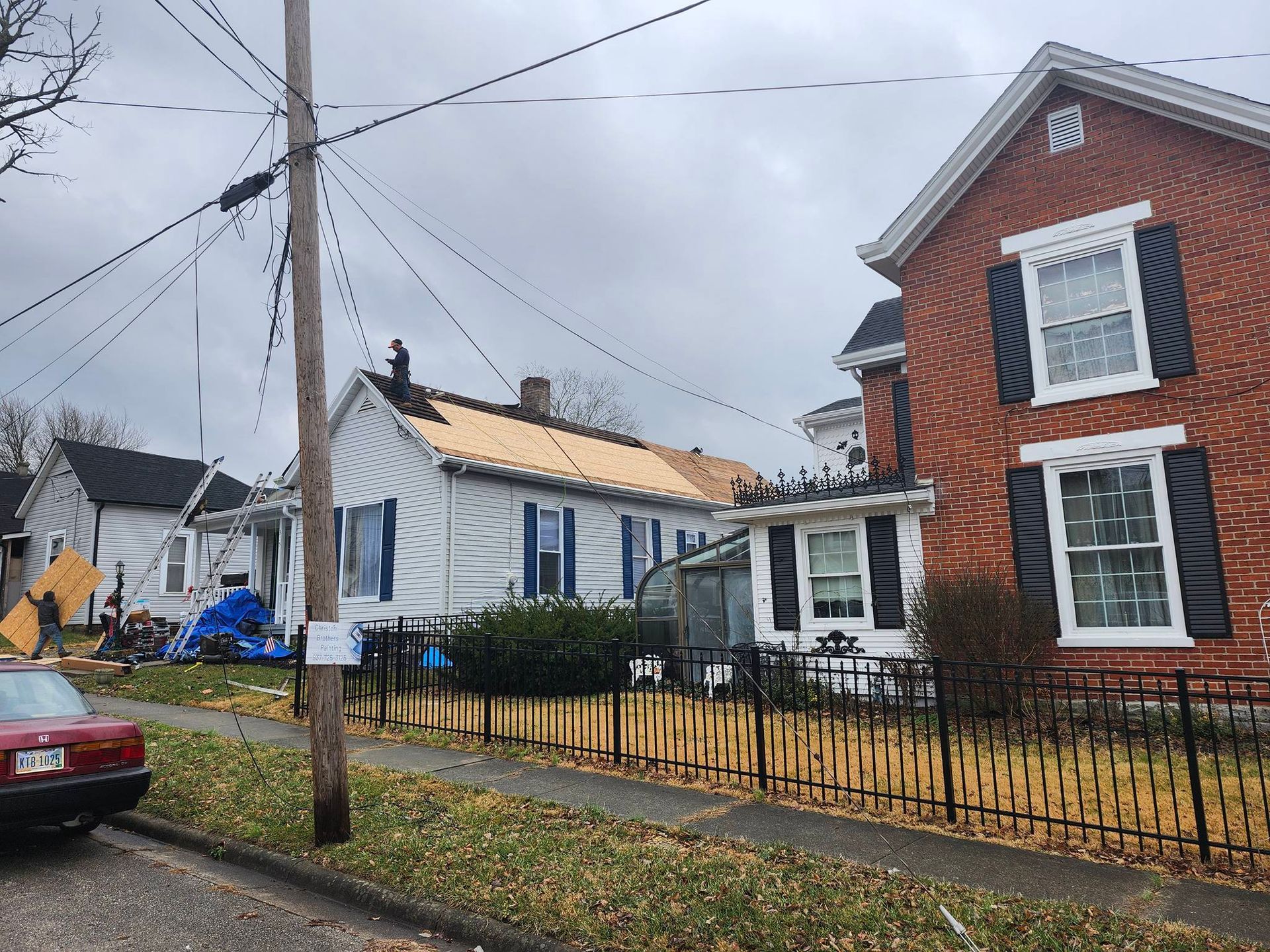 Rainy residential street with small houses, utility poles, and a black fence by a sidewalk.