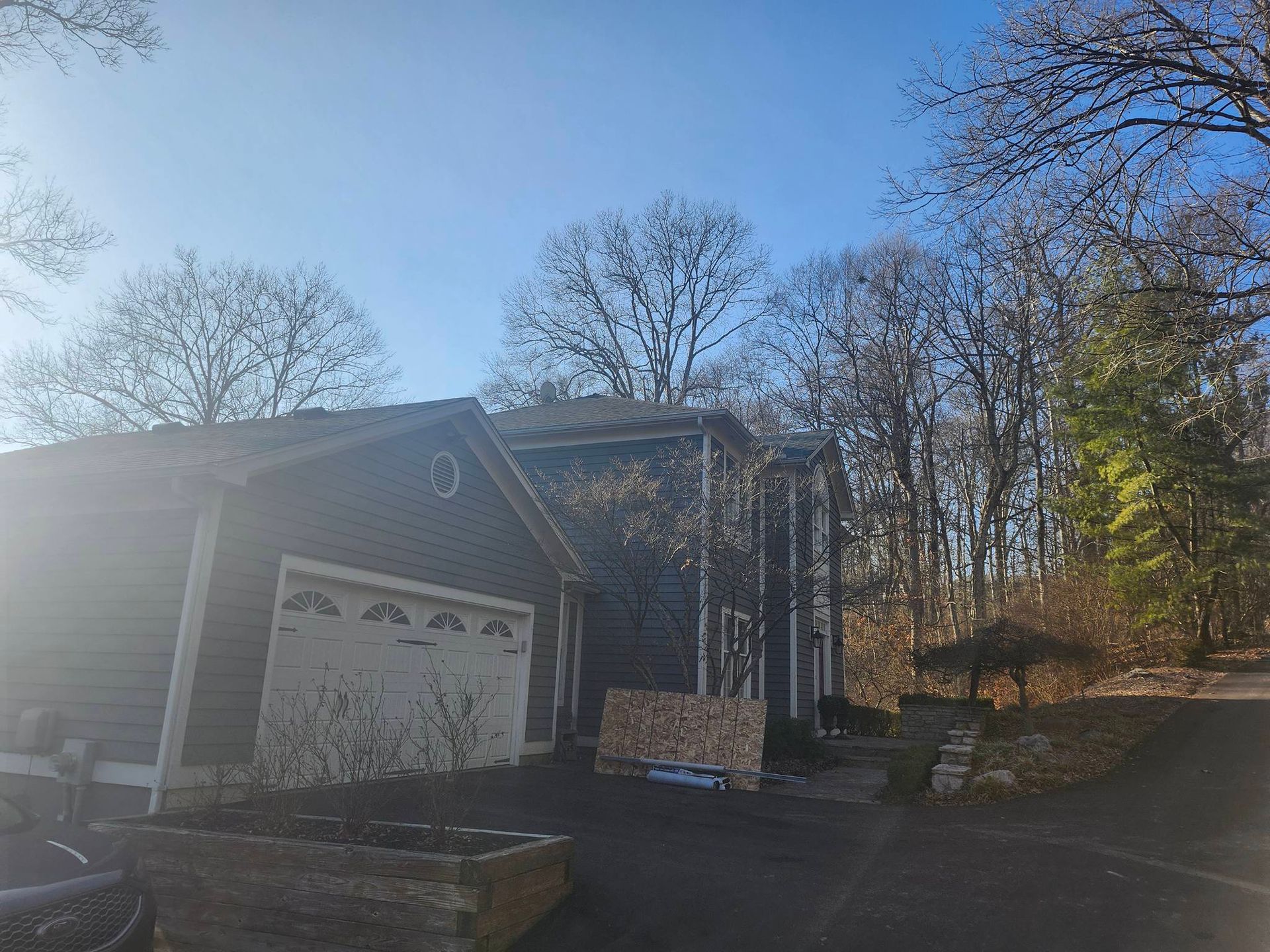 Suburban driveway with a garage, stacked firewood, and bare trees in winter sunlight