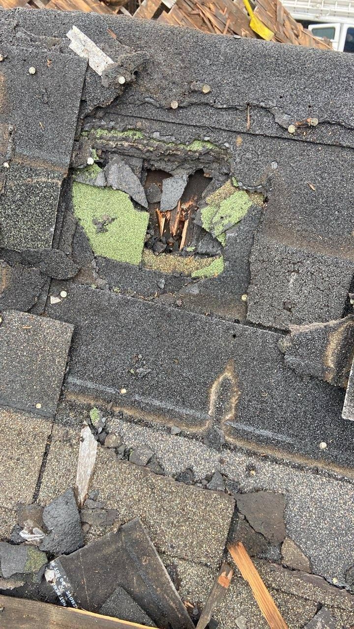 Damaged roof shingles with a hole exposing green underlayment and debris