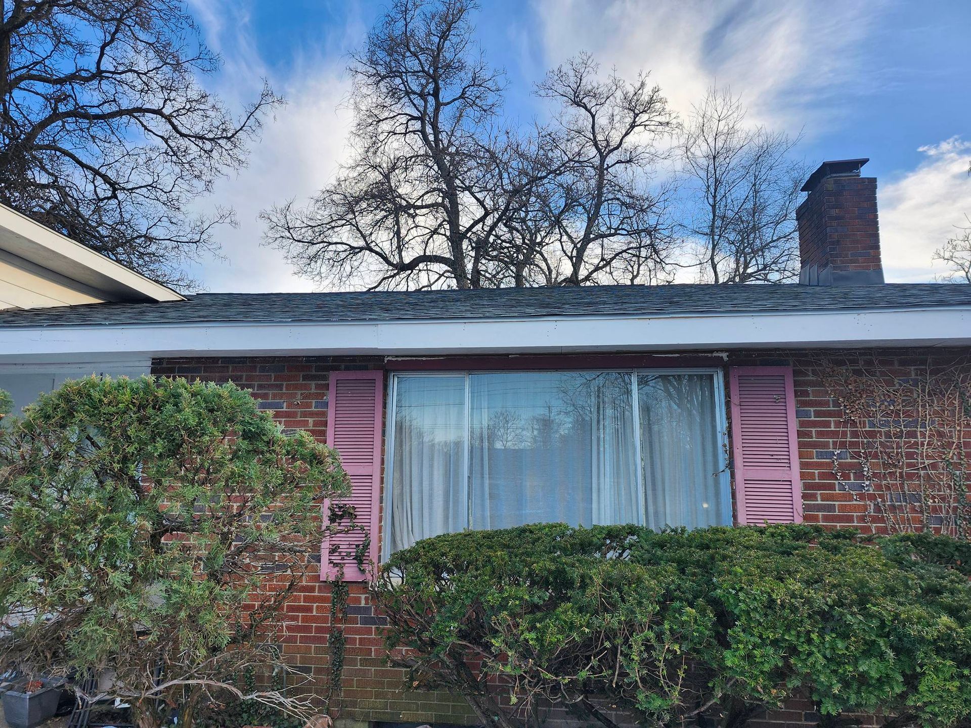 Brick house facade with pink shutters, large front window, hedges, and a chimney under a blue sky.