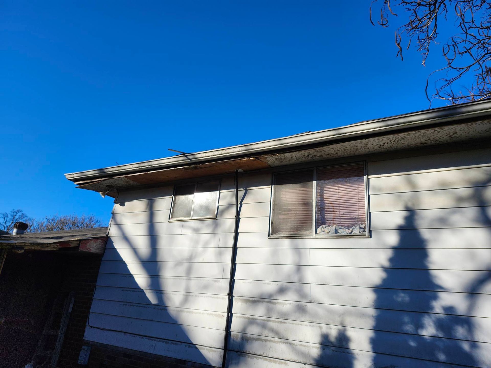 White house exterior with tan siding, a small window, and tree shadows under a clear blue sky