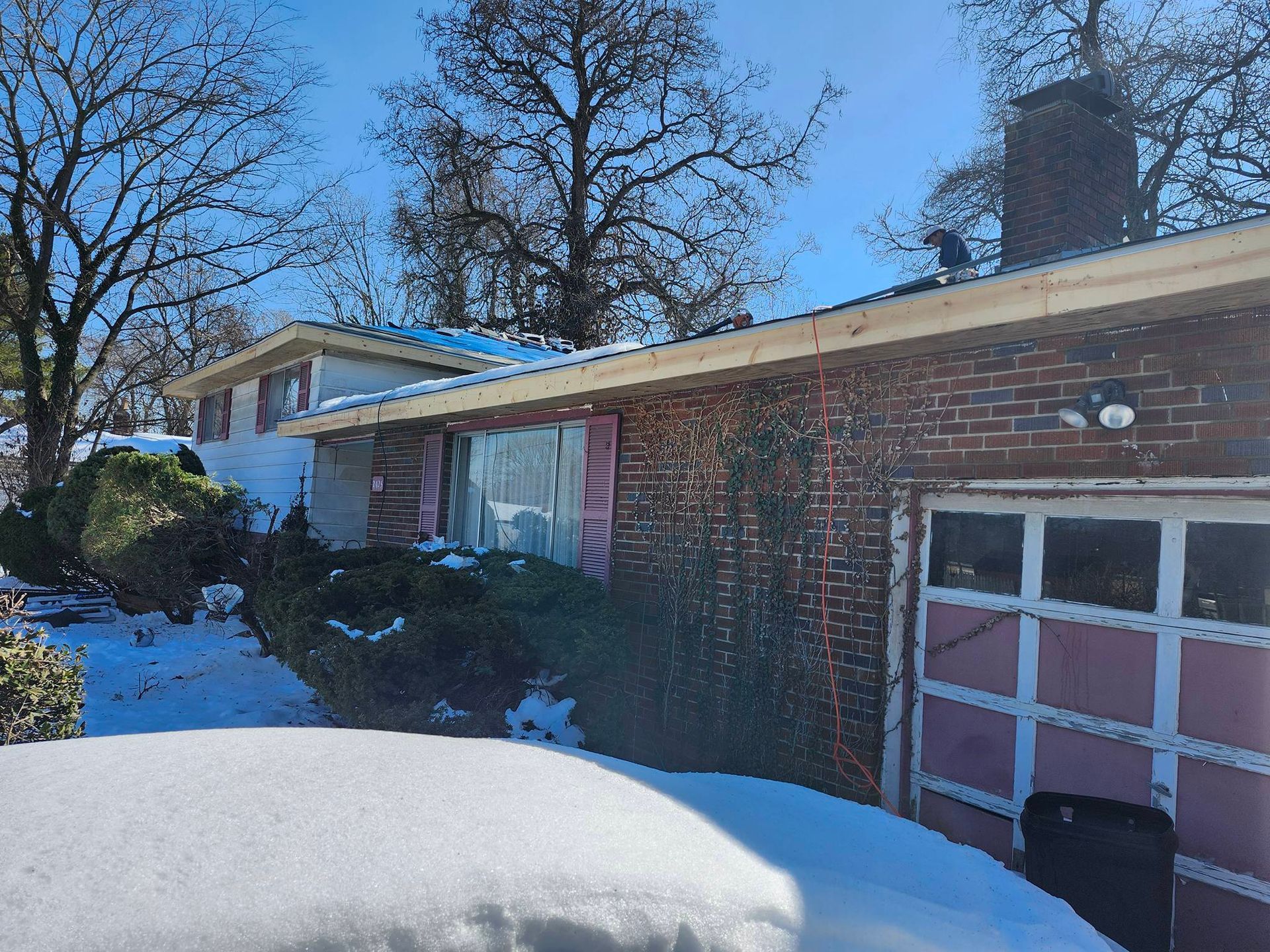 Snow-covered suburban brick house with garage and bare trees under a clear blue sky