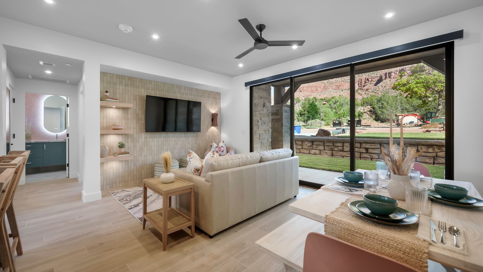 Bright living space with a large window, sofa, dining table, and a view of a grassy yard and red rock formations.