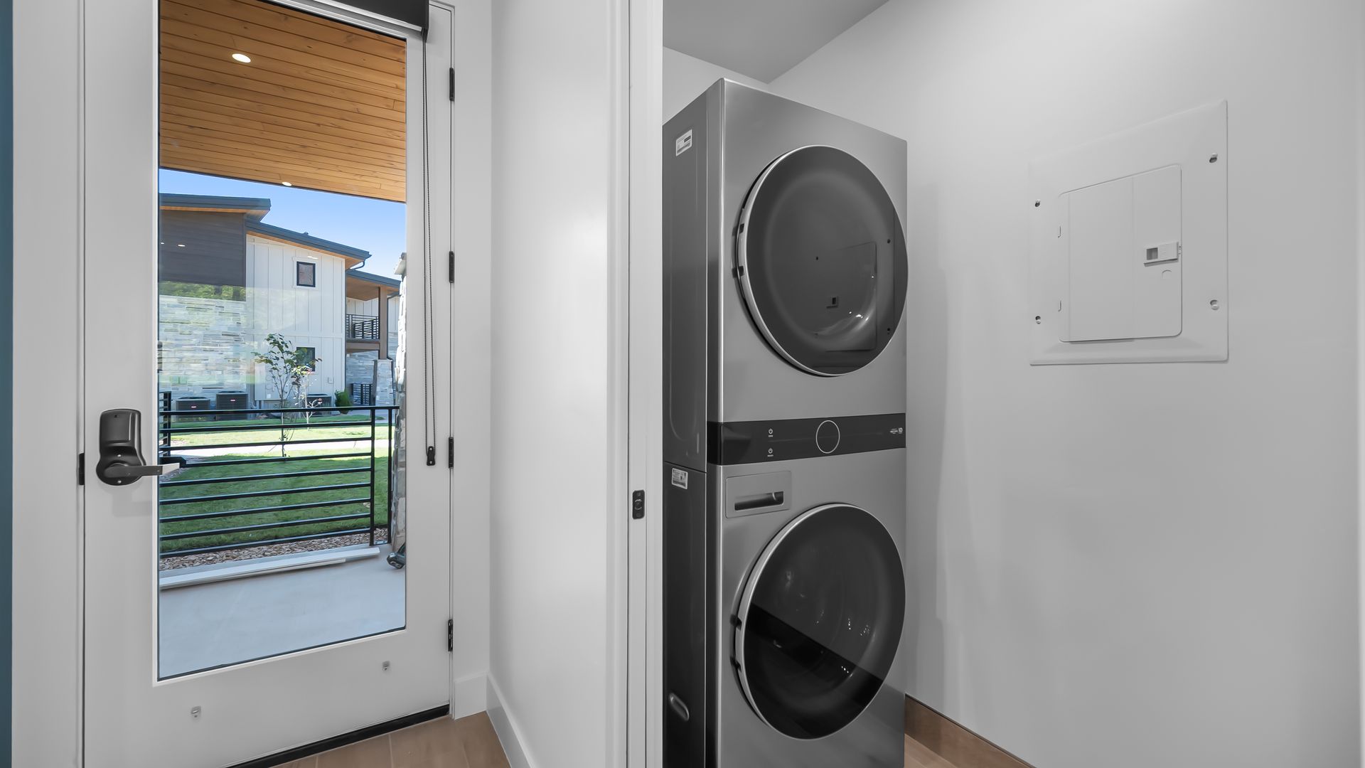 Stacked washer and dryer in a white laundry room, next to a glass door with an outdoor view.