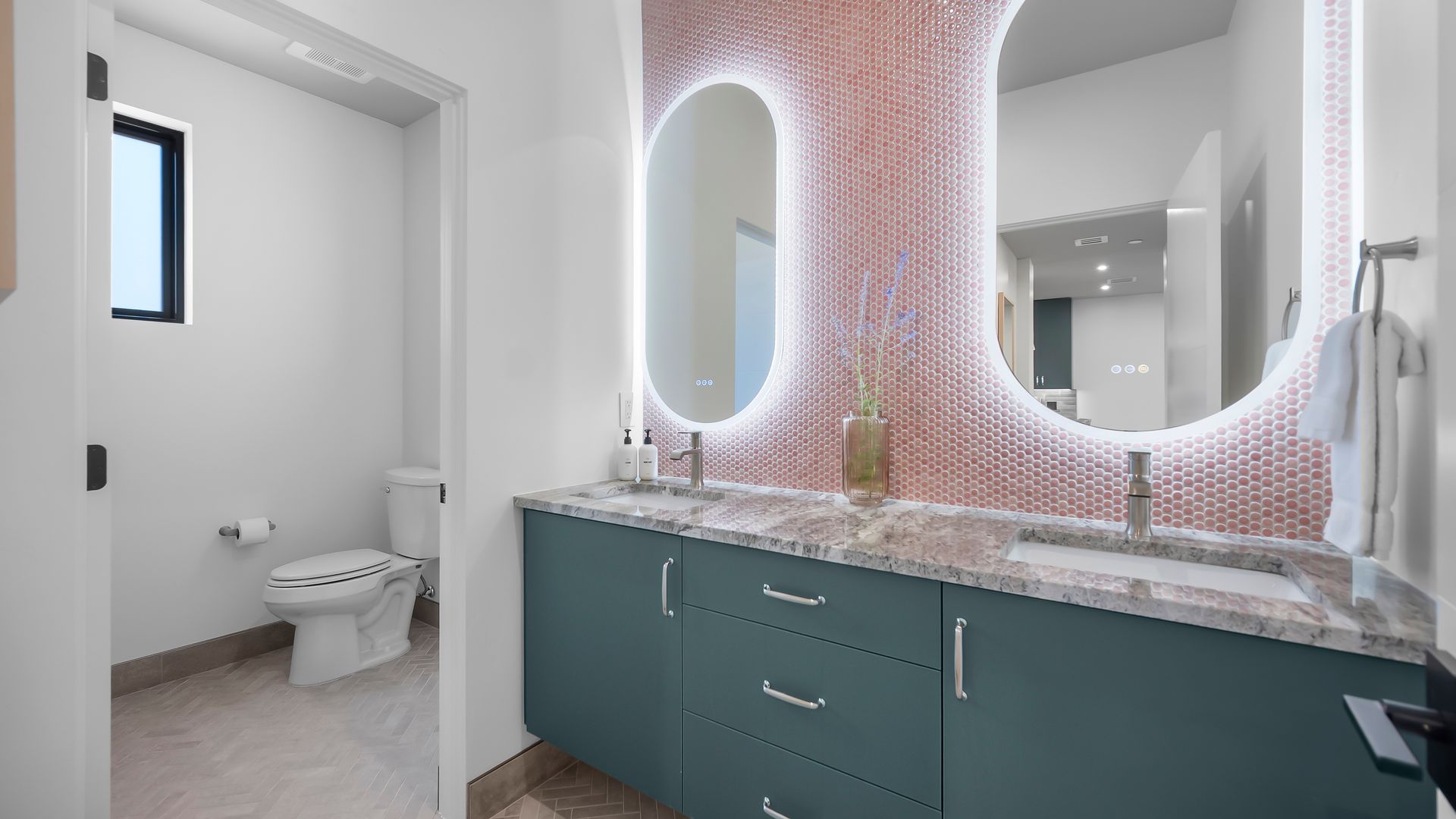 Bathroom with teal vanity, oval mirrors, pink tile wall, and a toilet.