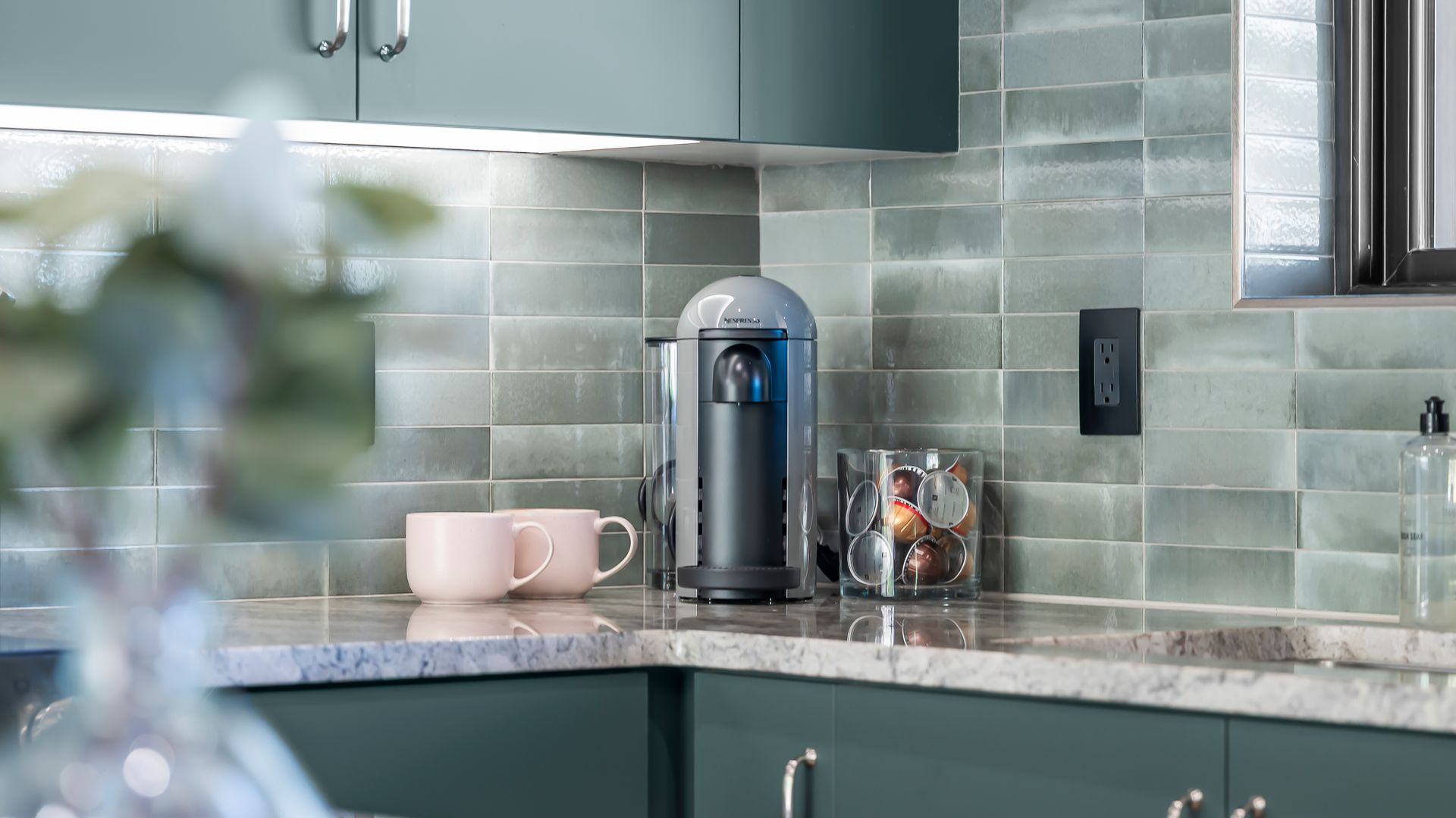 Kitchen counter with coffee maker, mugs, and black outlet on teal tiled backsplash.