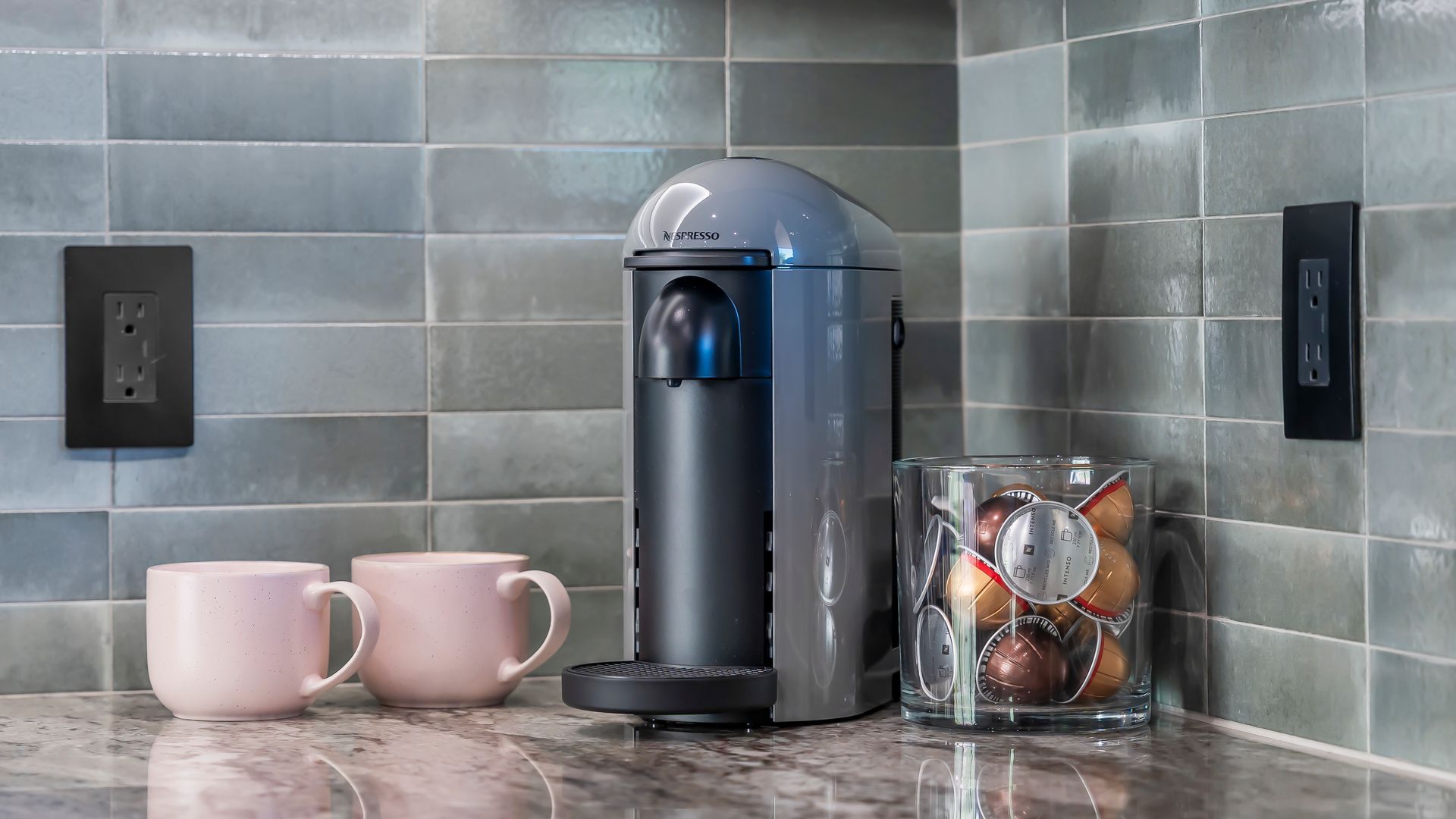 Gray coffee machine with pink mugs and coffee pods on a marble counter in a kitchen.