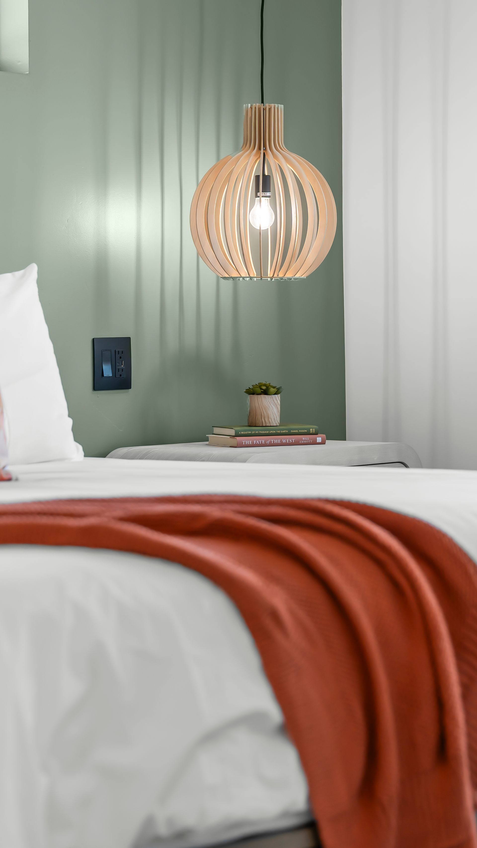 Bed with orange blanket, nightstand with pendant light and plant in front of sage green wall.